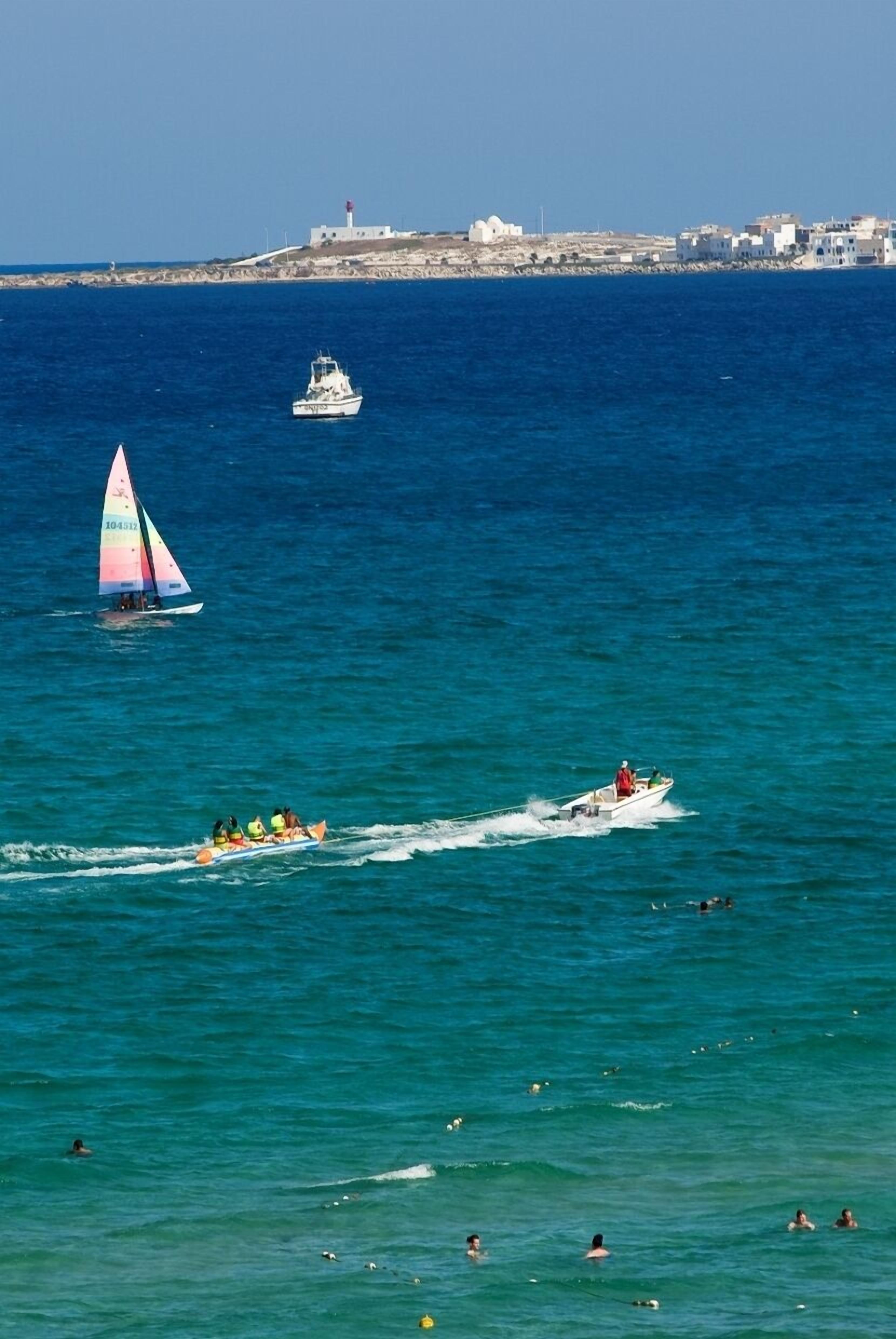 A coastal scene with a sailboat, a motorboat, and swimmers in the water.
