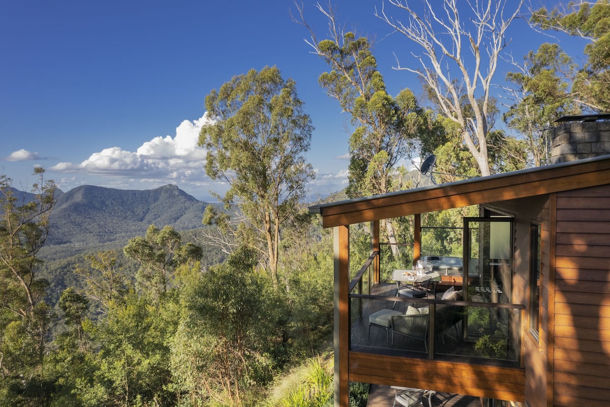 A wooden cabin with a deck, large windows, and a view of mountains and trees.