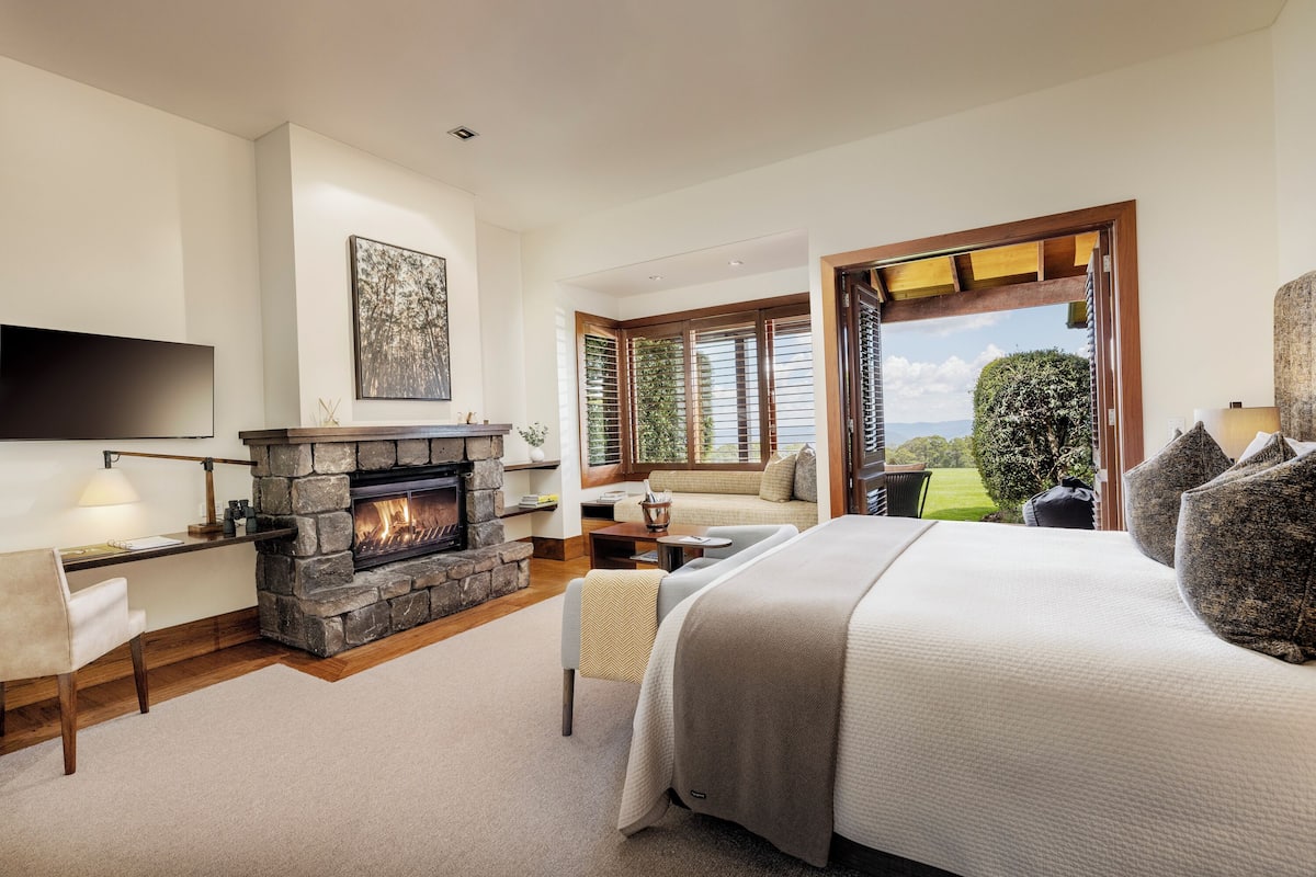 A modern bedroom with a stone fireplace, a large bed, a flat-screen TV, and a view of the outdoors through a large window.