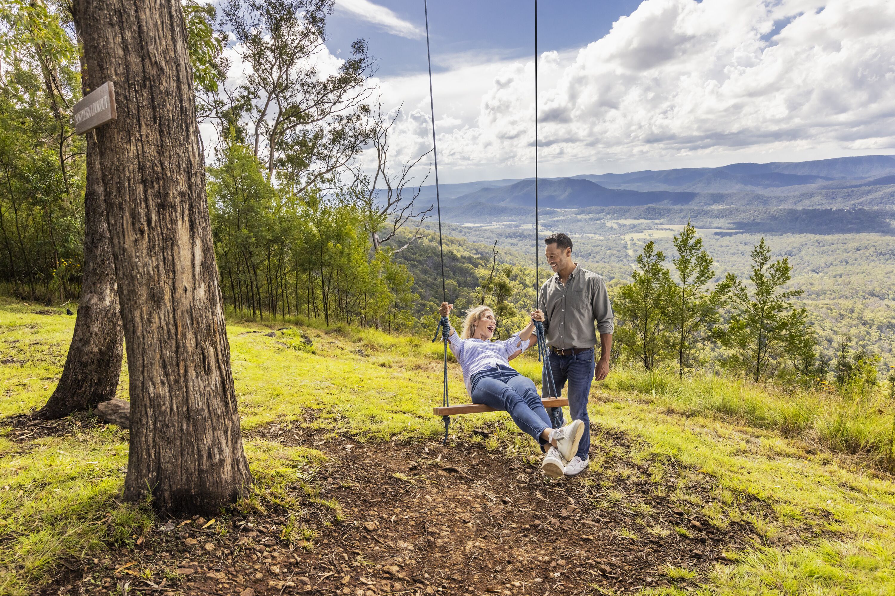A person swinging on a bench in a natural setting with a view of distant mountains.