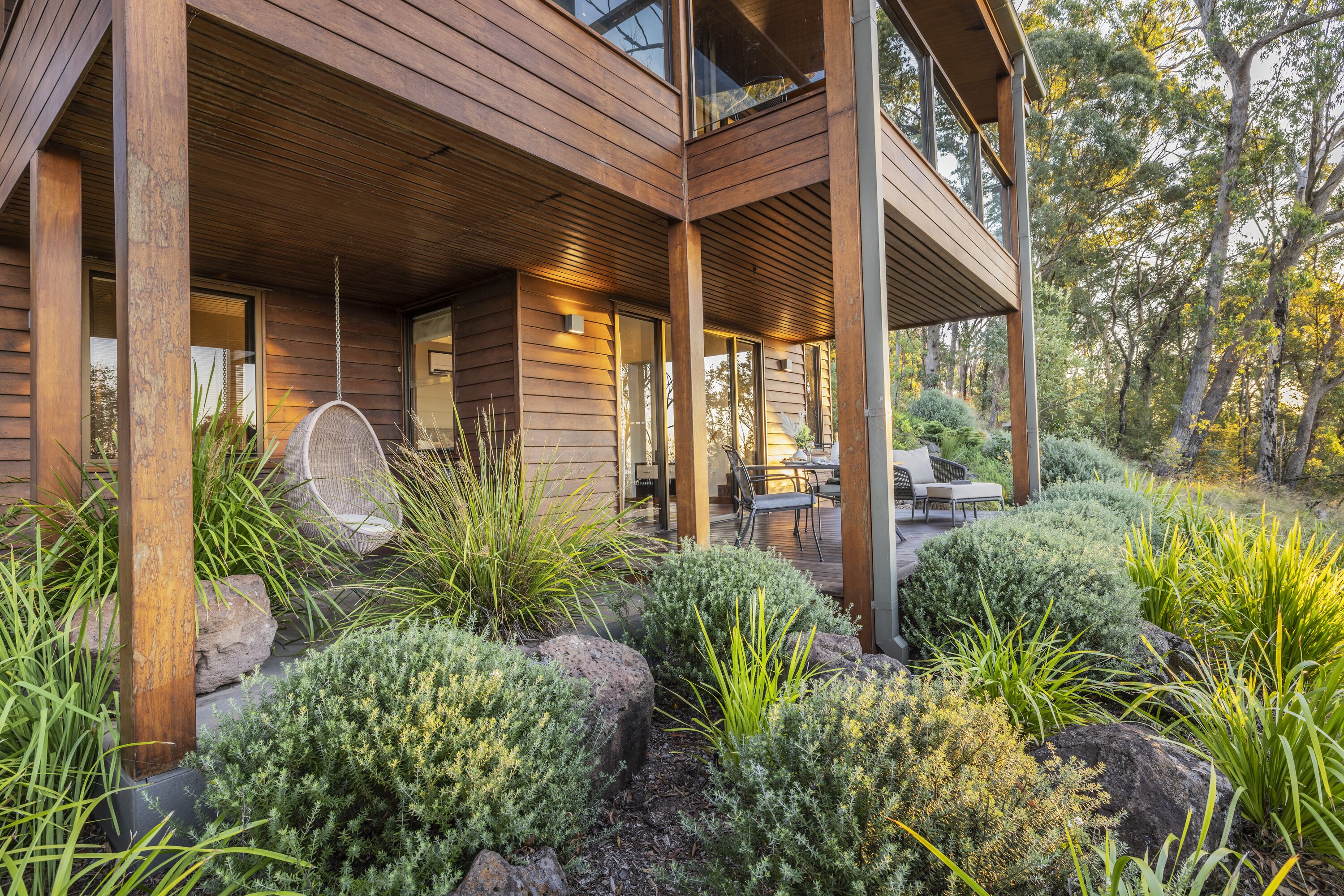 Una casa moderna de madera con un porche cubierto, mobiliario exterior y un jardín con diversas plantas y rocas.