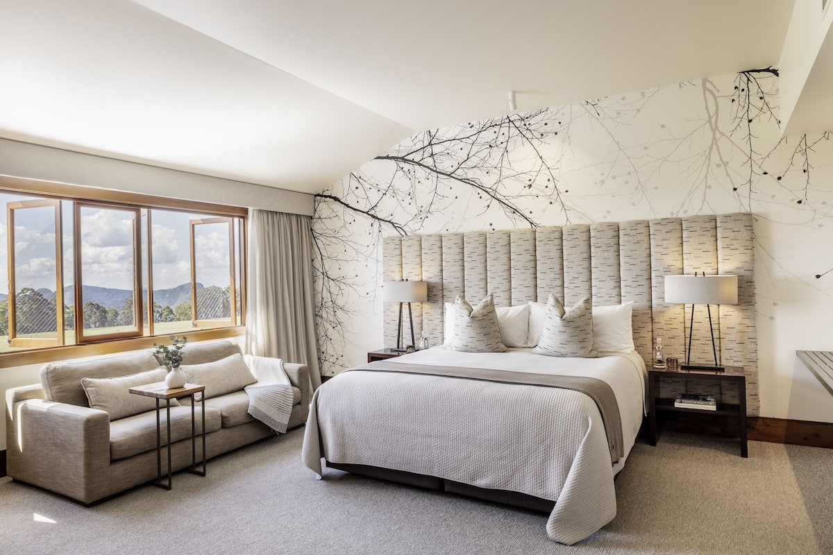 A modern hotel room with a large bed, a sofa, and a view of the mountains through large windows.