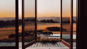 An open window with a view of a sunset over a field, a wooden deck, and a small garden area. - Spicers Peak Lodge (Swanfels)