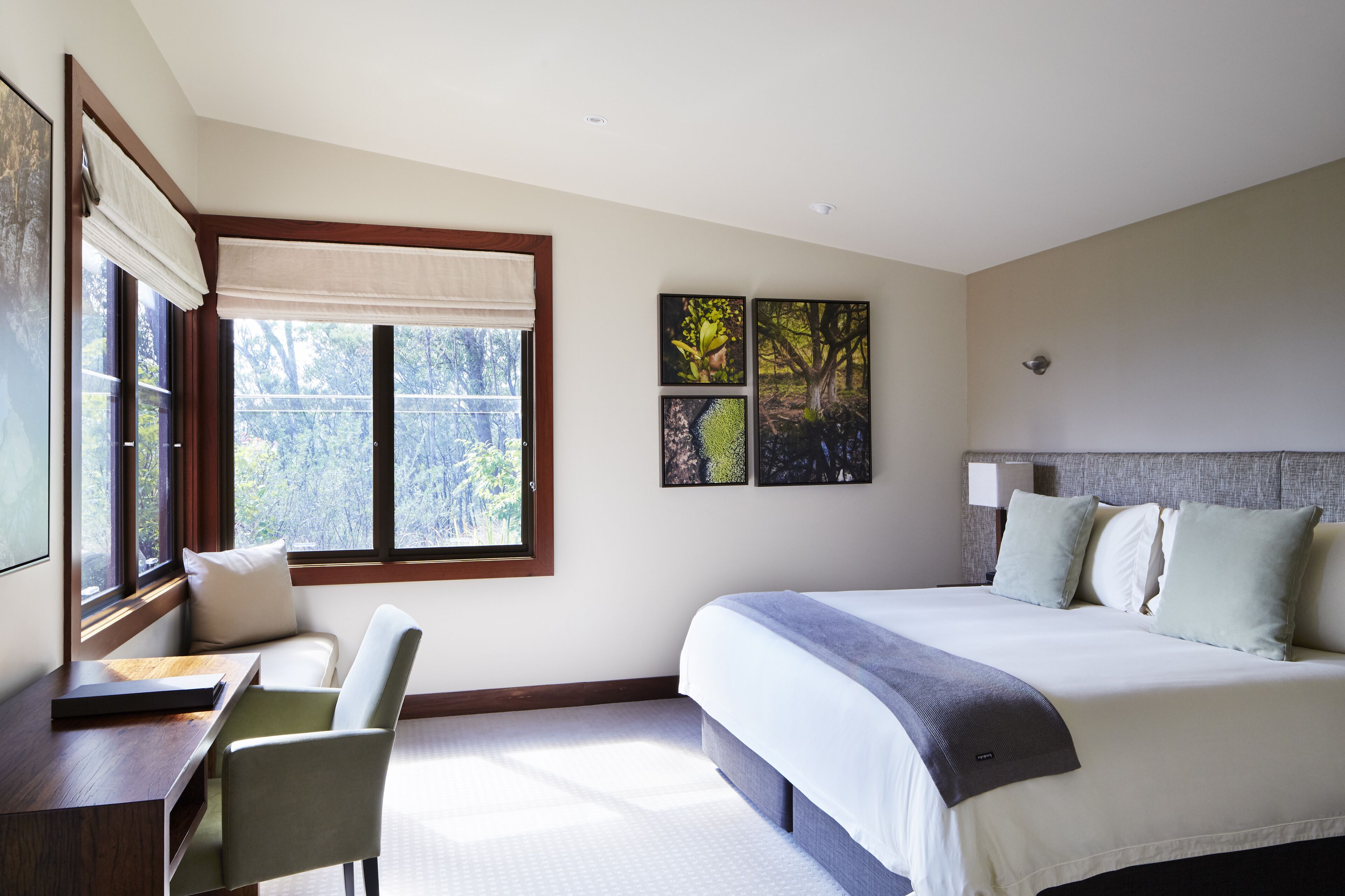 A bedroom with a bed, a desk, and a chair. There are two windows with curtains, and artwork on the wall.