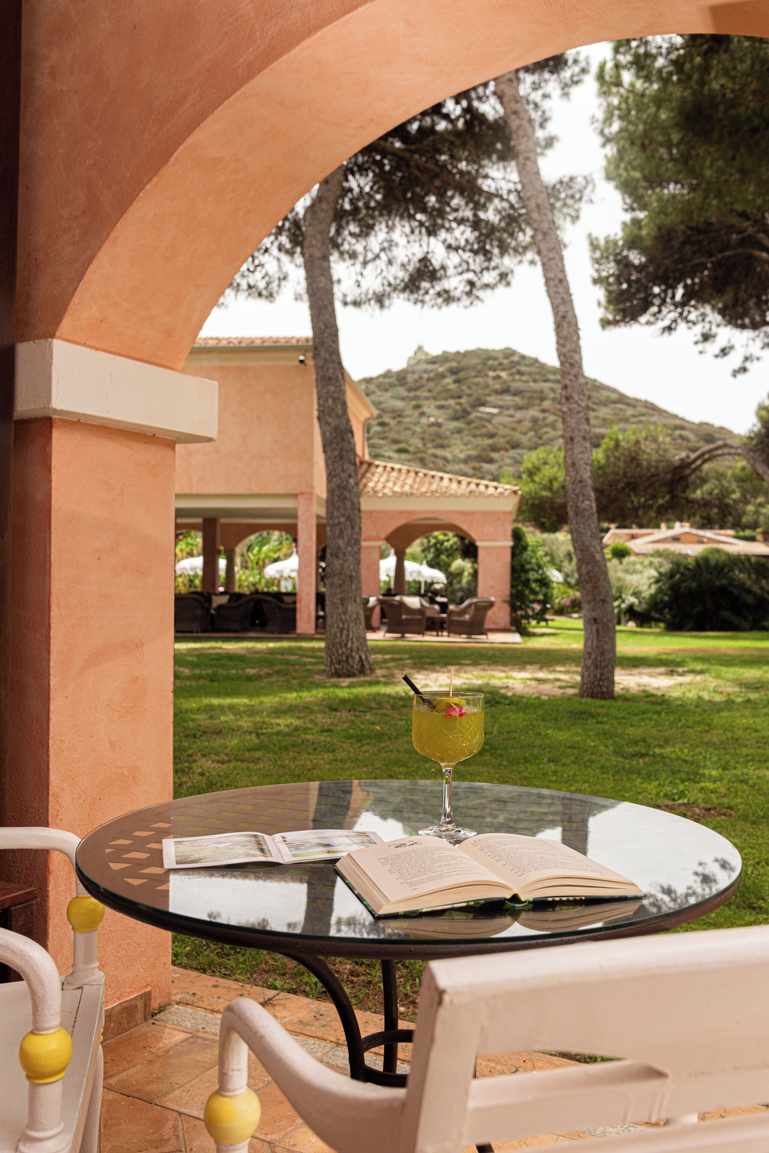 A glass table with an open book and a drink, set in a covered outdoor area with chairs and a view of a building and trees.