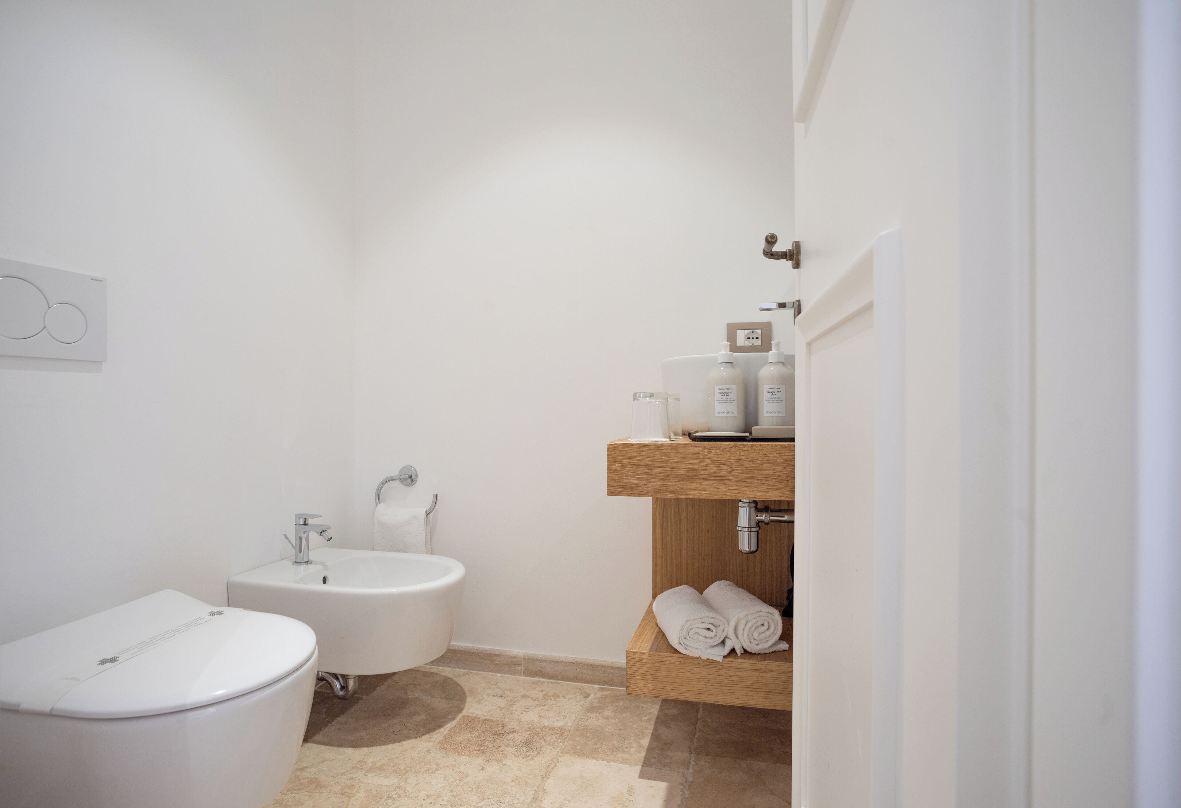 Une salle de bain moderne équipée de toilettes, d’un bidet et d’une étagère en bois où sont rangées les serviettes.