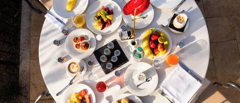 Une table de salle à manger ronde, sur laquelle on trouve divers plats, notamment des fruits, des pâtisseries, et un parasol rouge.