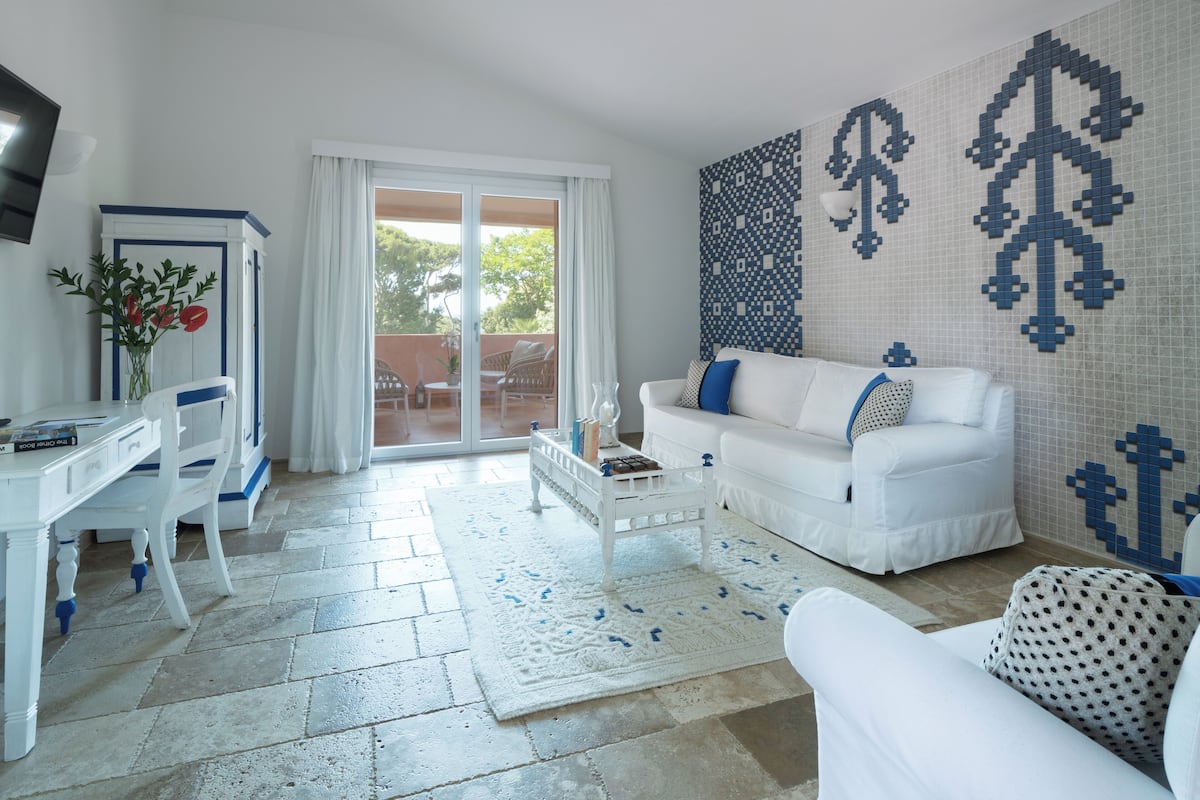 Een moderne woonkamer met een witte bank, een witte salontafel en een tapijt met een patroon. Er staat een witte kast met een bloevas, en een groot raam met uitzicht op de omgeving.