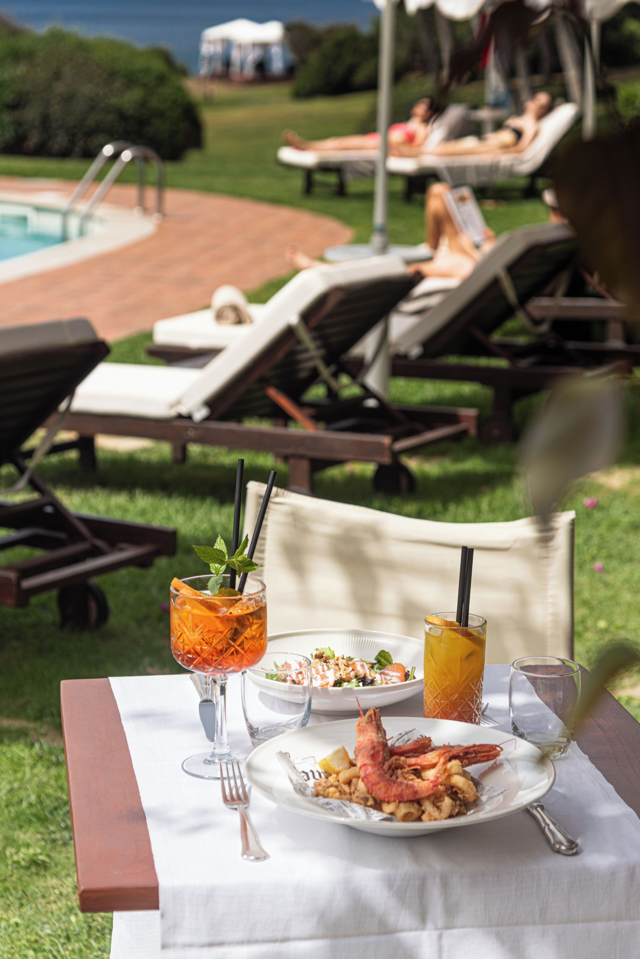 Une table à manger avec un repas, deux boissons, et une vue sur une piscine et des chaises longues.