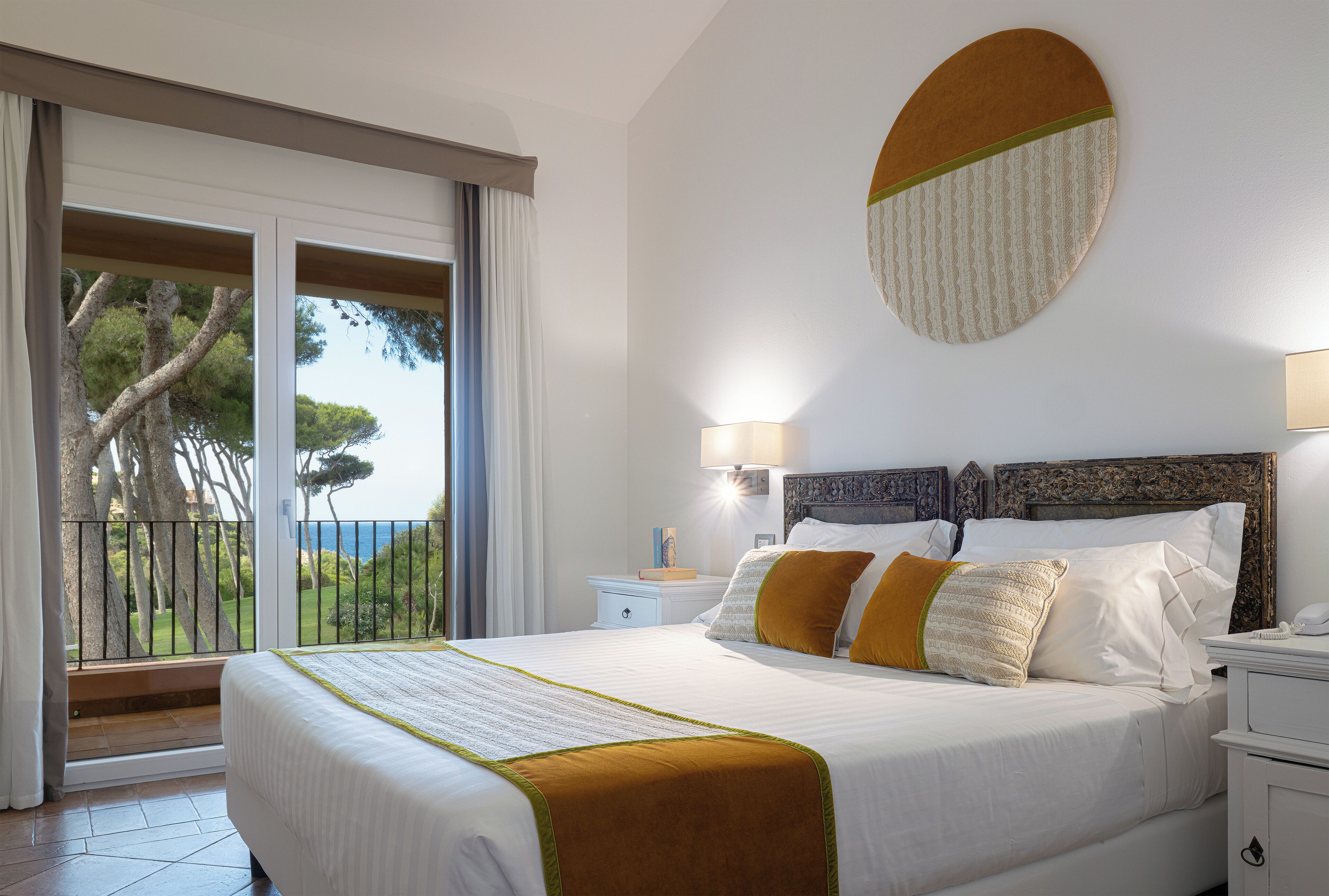 Ein Hotelzimmer mit einem großen Bett, einem Balkon mit Blick auf Bäume und Meer, einer runden Wanddekoration und einem Nachttisch mit Lampe.