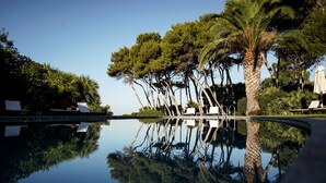 Un área de piscina tranquila con sillones de descanso y palmeras, que se reflejan en el agua.