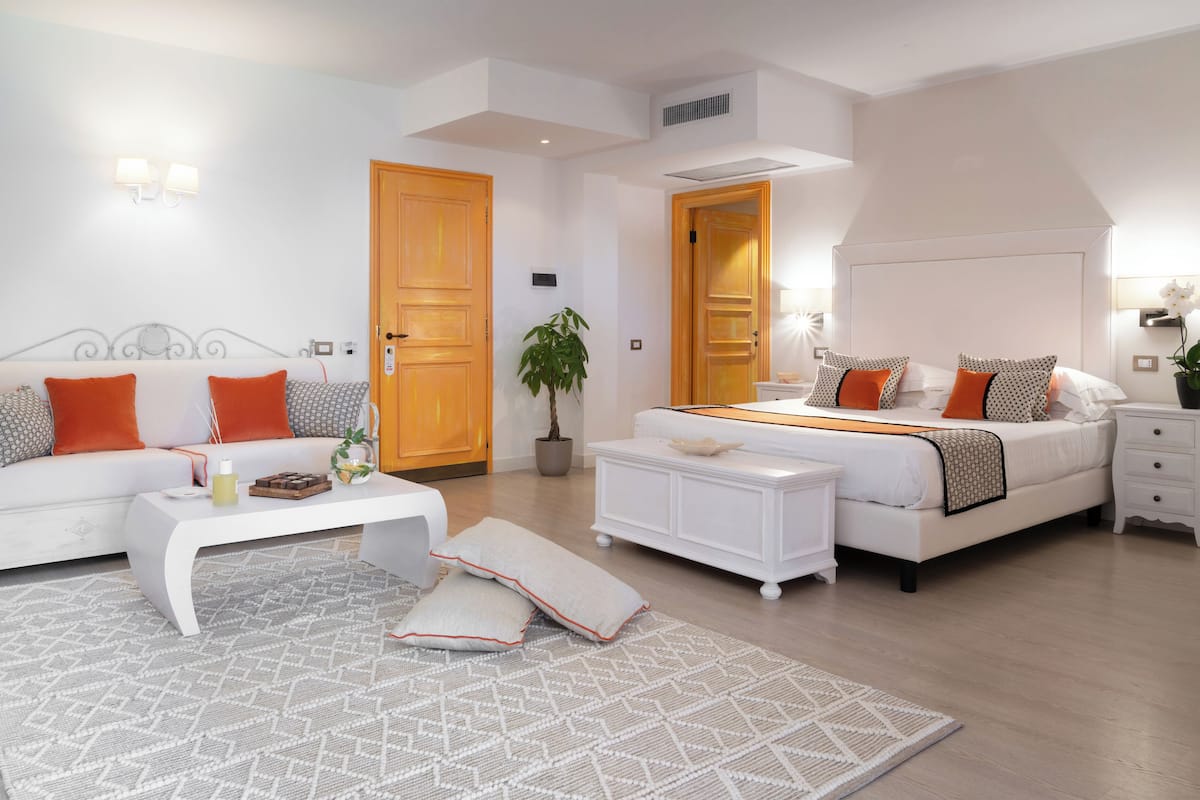 Een moderne slaapkamer met een witte bank, een wit bed en een witte nachtkastje. Op de vloer ligt een kamerkleed met een patroon en er staat een potplant in de hoek.