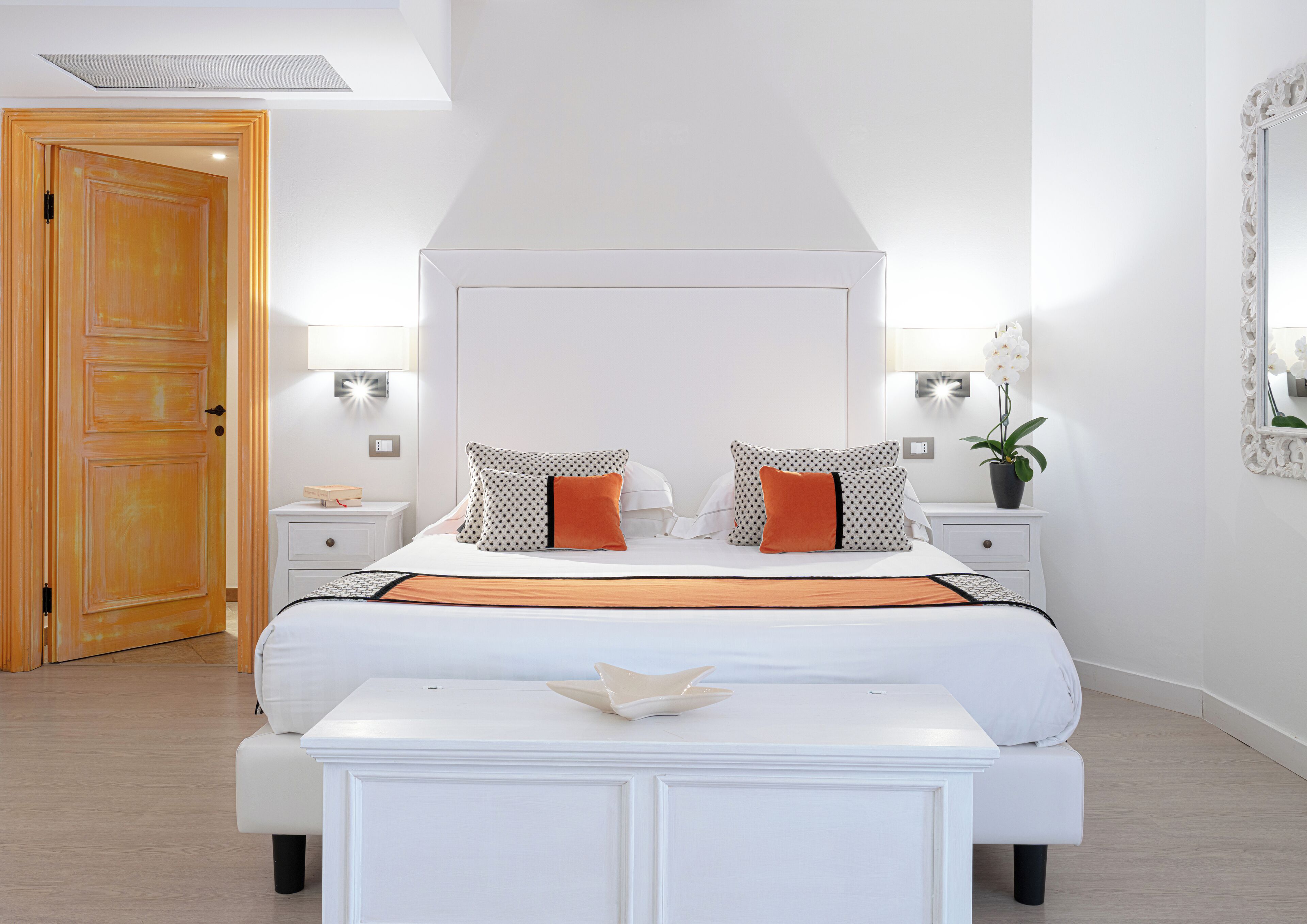 Un dormitorio con una cama grande, dos mesitas de noche, un taburete, una puerta de madera y un espejo en la pared.