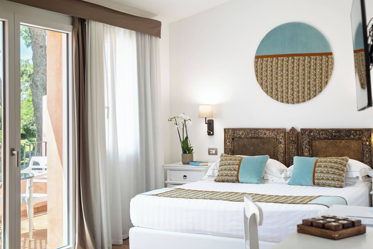 Een hotelkamer met een bed, nachtkastje, lamp en uitzicht op de omgeving.