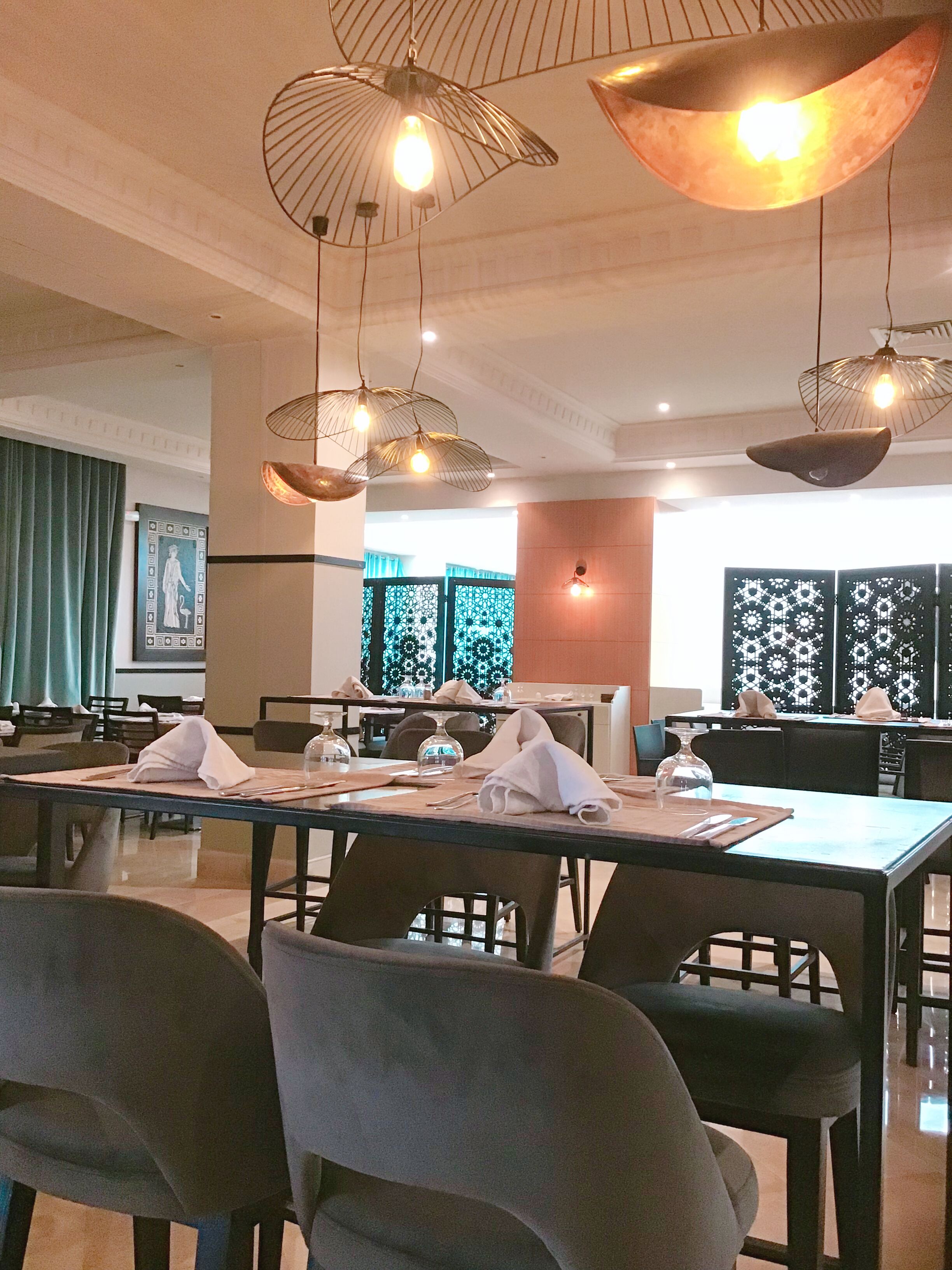 Restaurante com mesas preparadas para refeições, luminárias pendentes e vista para a área de jantar através de um separador.
