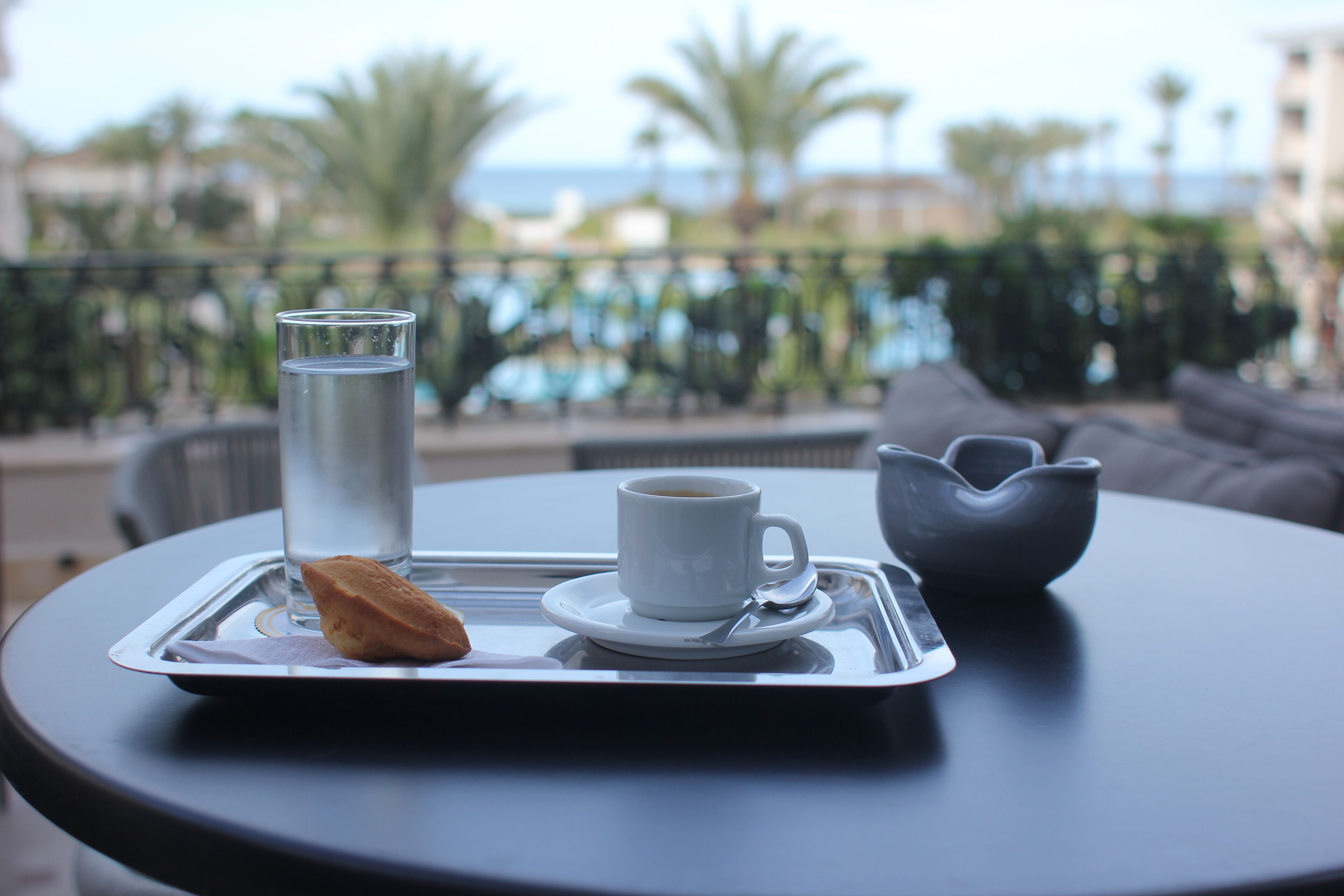 Una bandeja con una taza de café, un vaso de agua y un pastel sobre una mesa con vistas a una piscina y palmeras.