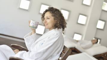 Eine Person in einem weißen Bademantel sitzt in einem Sessel mit einer Tasse in einem Raum mit mehreren Betten und modernem, minimalistischen Design.