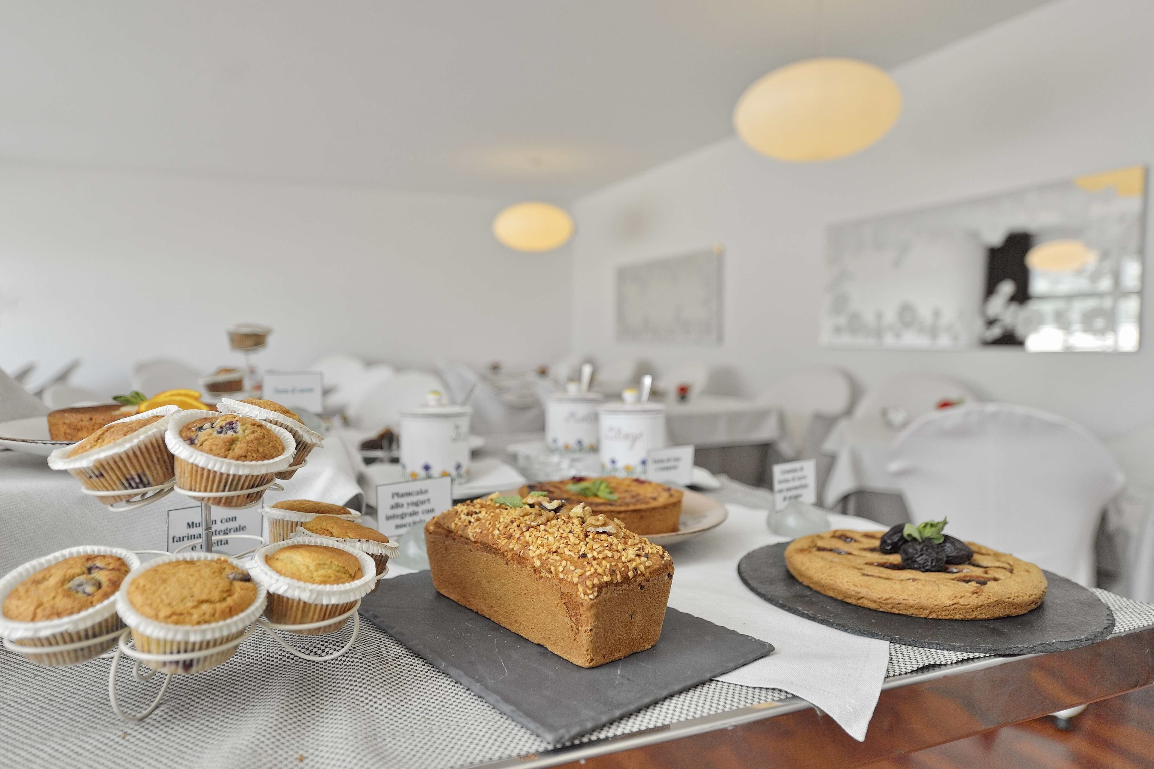 A buffet table with various baked goods, including muffins and a loaf of bread, in a modern, well-lit room with white decor and artwork on the walls.