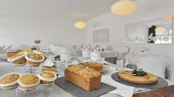 A buffet table with various baked goods, including muffins and a loaf of bread, in a modern, well-lit room with white decor and artwork on the walls.