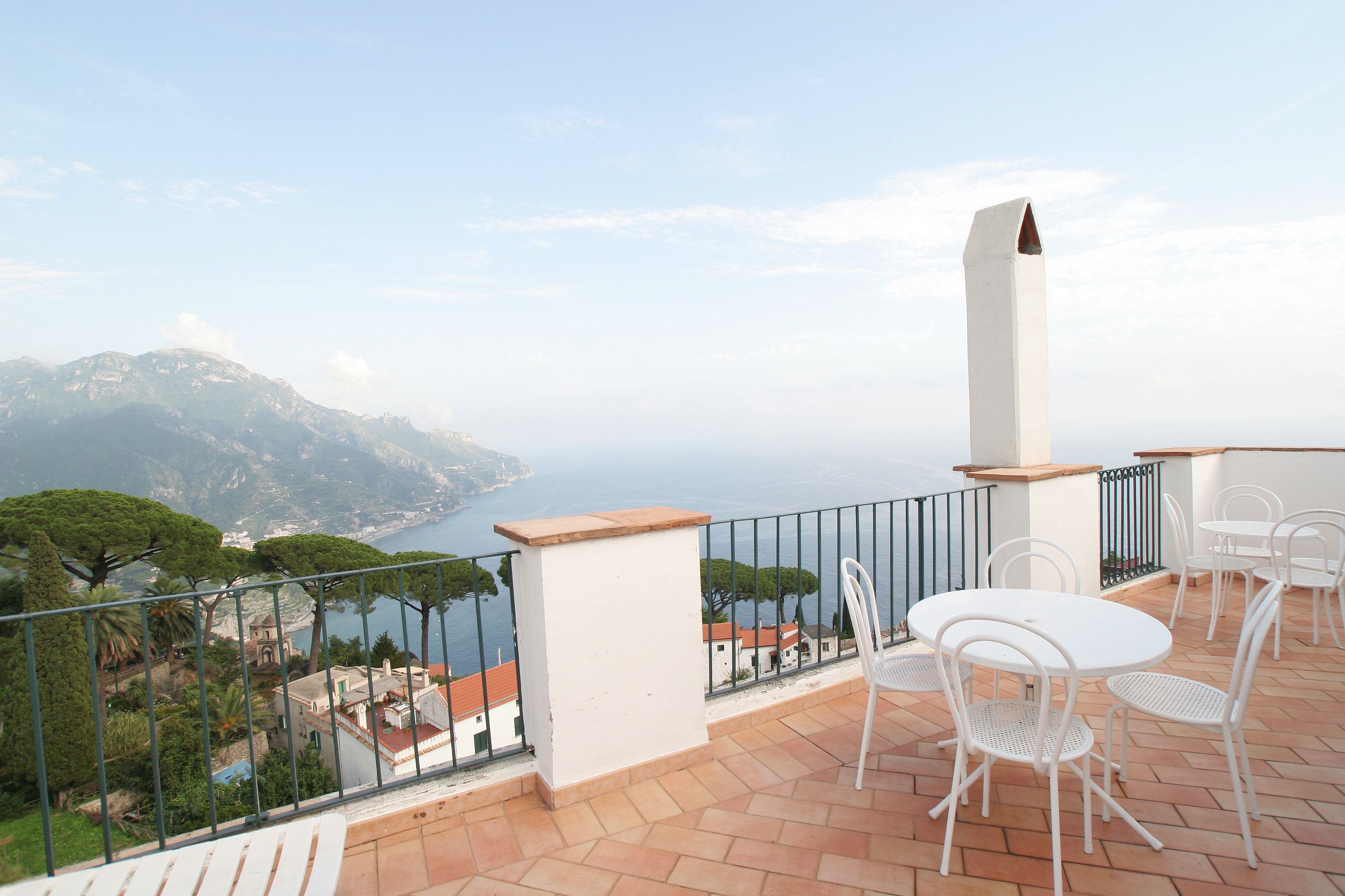 A terrace with white chairs and a table, overlooking a coastal landscape with buildings and trees.