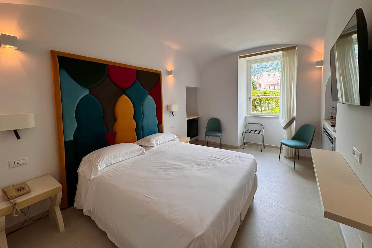 A hotel room with a large bed, a desk, a chair, a TV, and a window with a view of buildings.