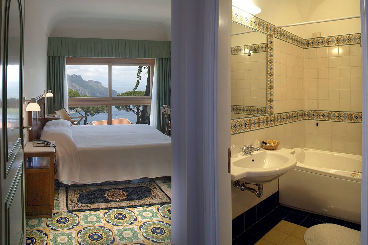 A hotel room with a bed, a desk, a telephone, a window with a view of mountains, and a bathroom with a bathtub and sink.