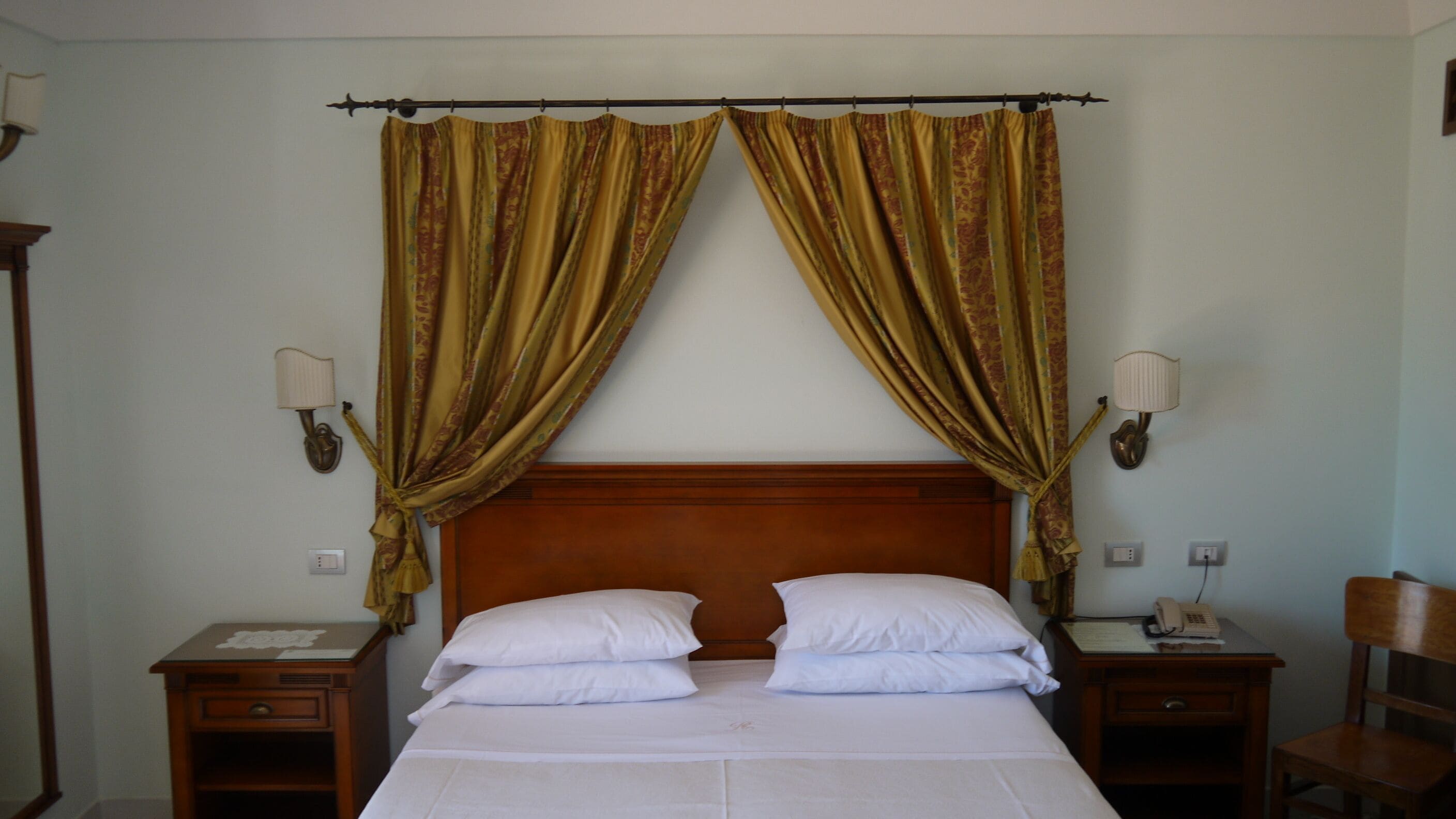 A hotel room with a bed, two bedside tables, a chair, and a curtain with a pattern.