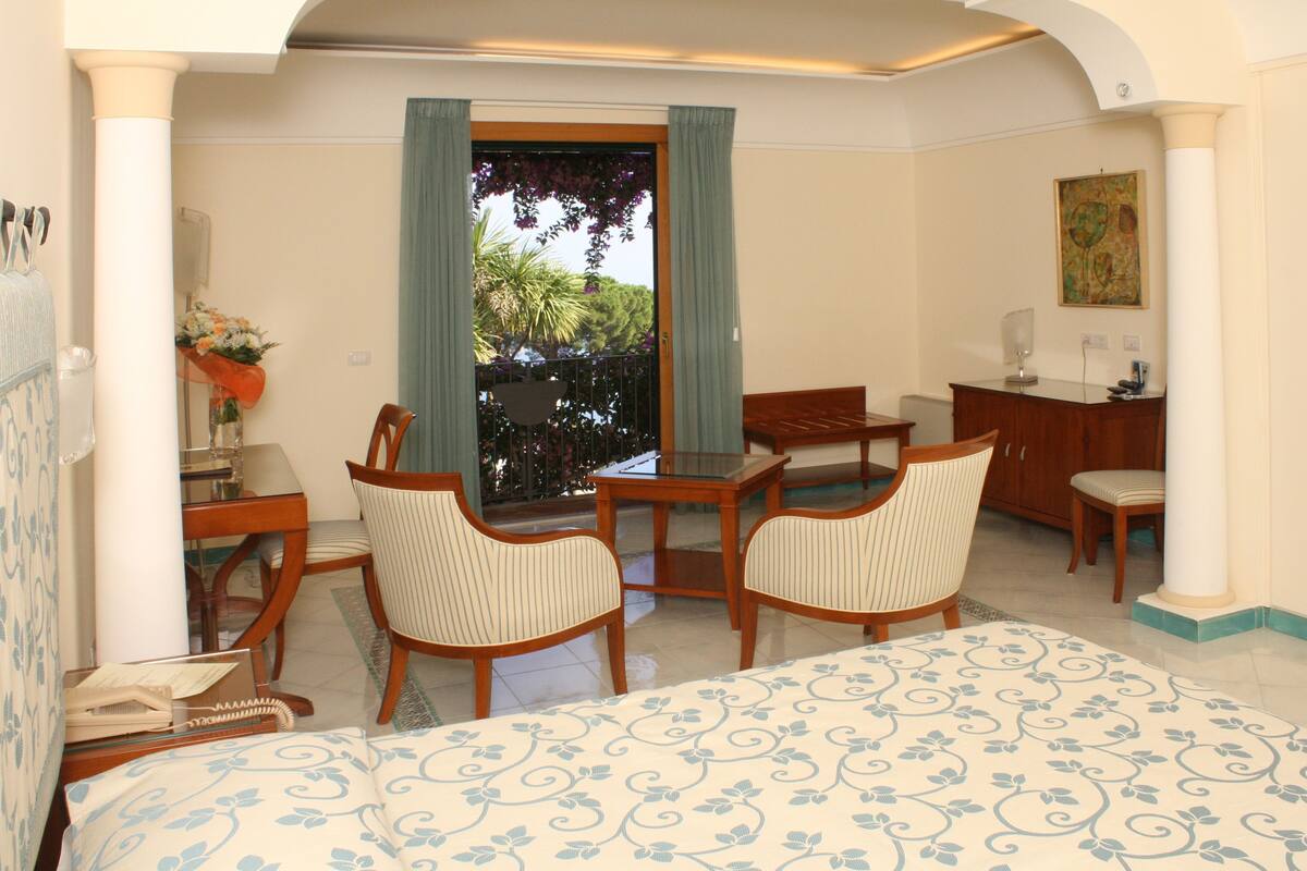 A hotel room with a bed, two wooden chairs, a small table, a desk, a desk chair, a nightstand, and a window with curtains.