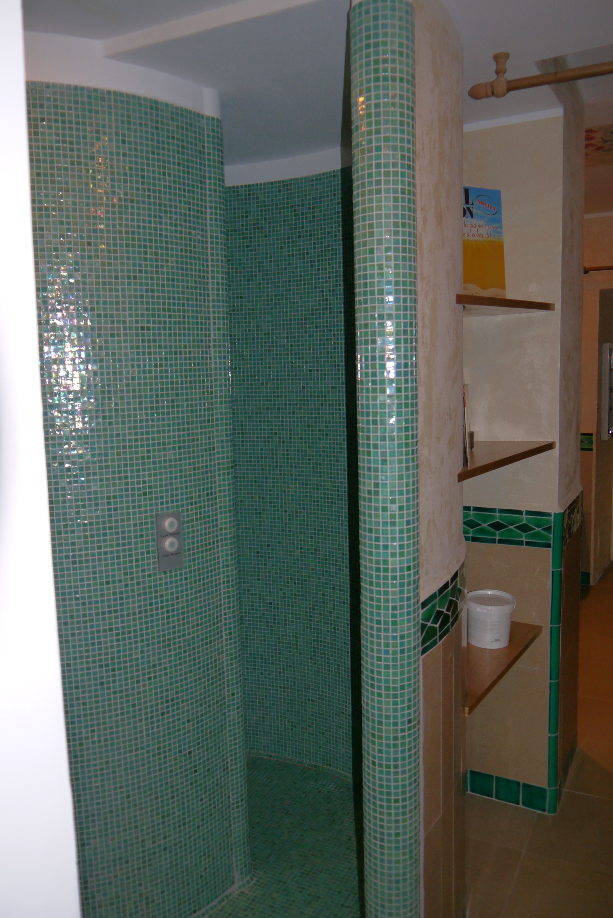 Une salle de bain avec des carreaux de mosaïque verts, un espace douche et une étagère en bois sur laquelle est posée une serviette.
