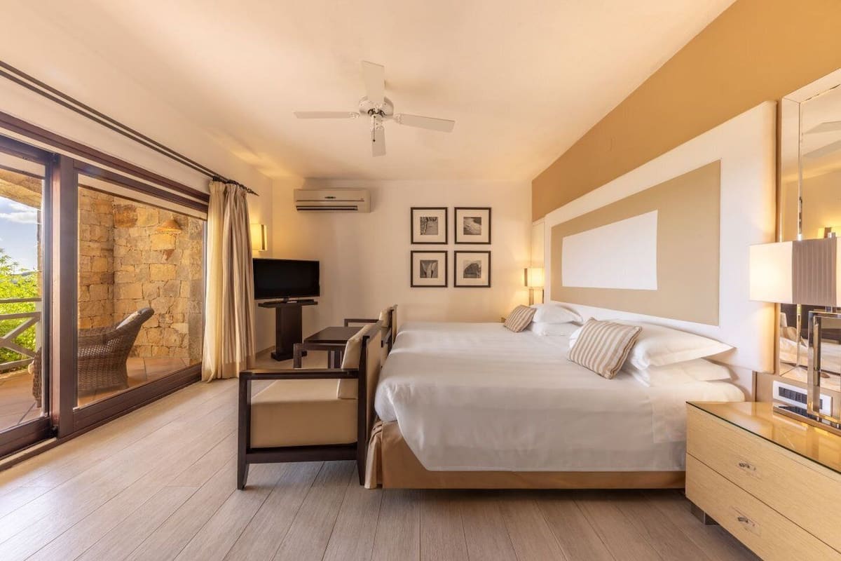 Ein Hotelzimmer mit einem großen Bett, einem Schreibtisch mit Stuhl, einem Fernseher und einem Balkon mit Korbmöbeln.