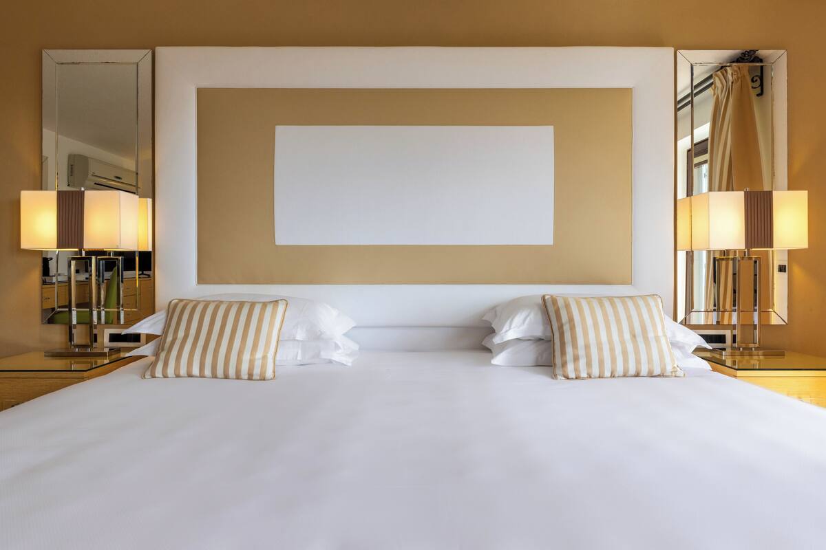 Ein Hotelzimmer mit einem großen Bett, zwei Nachttischlampen, einem Spiegel und einer leeren Wand über dem Bett.