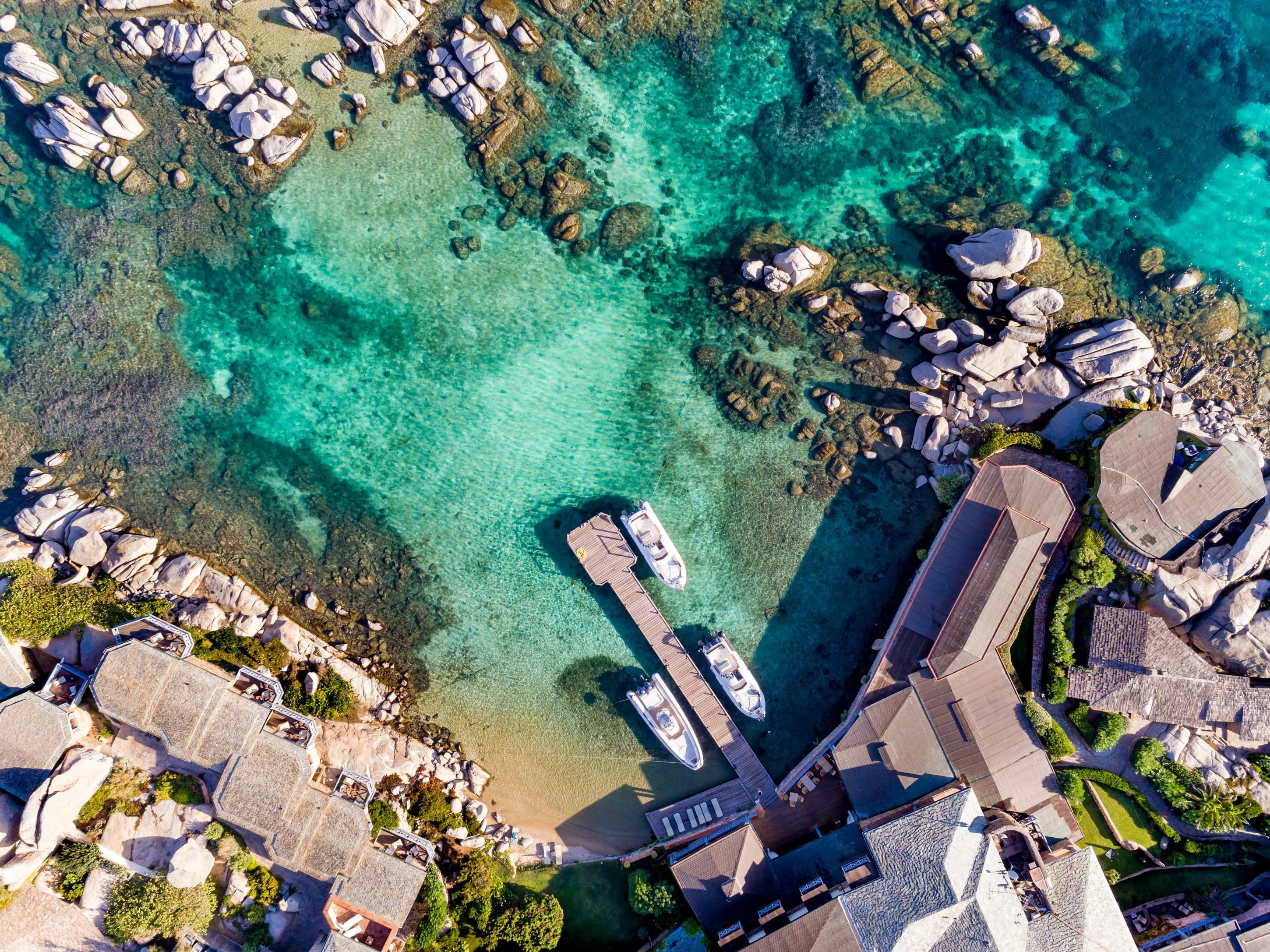 An aerial view of a coastal area with clear turquoise waters, a dock with boats, and a cluster of buildings with distinct architectural styles.