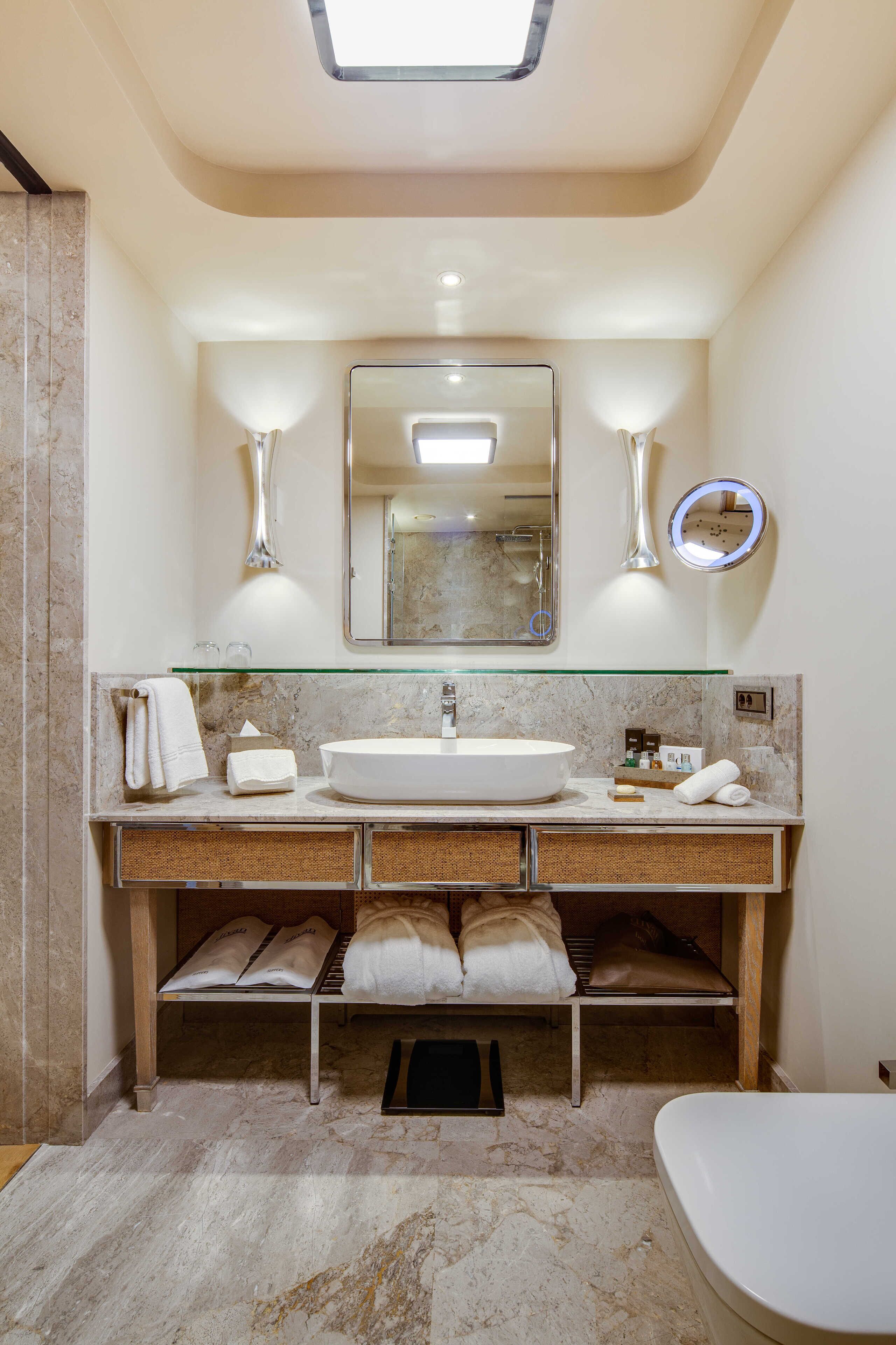 Un baño moderno con un espejo grande, un espejo redondo, un estante de vidrio sobre el lavabo y un inodoro.