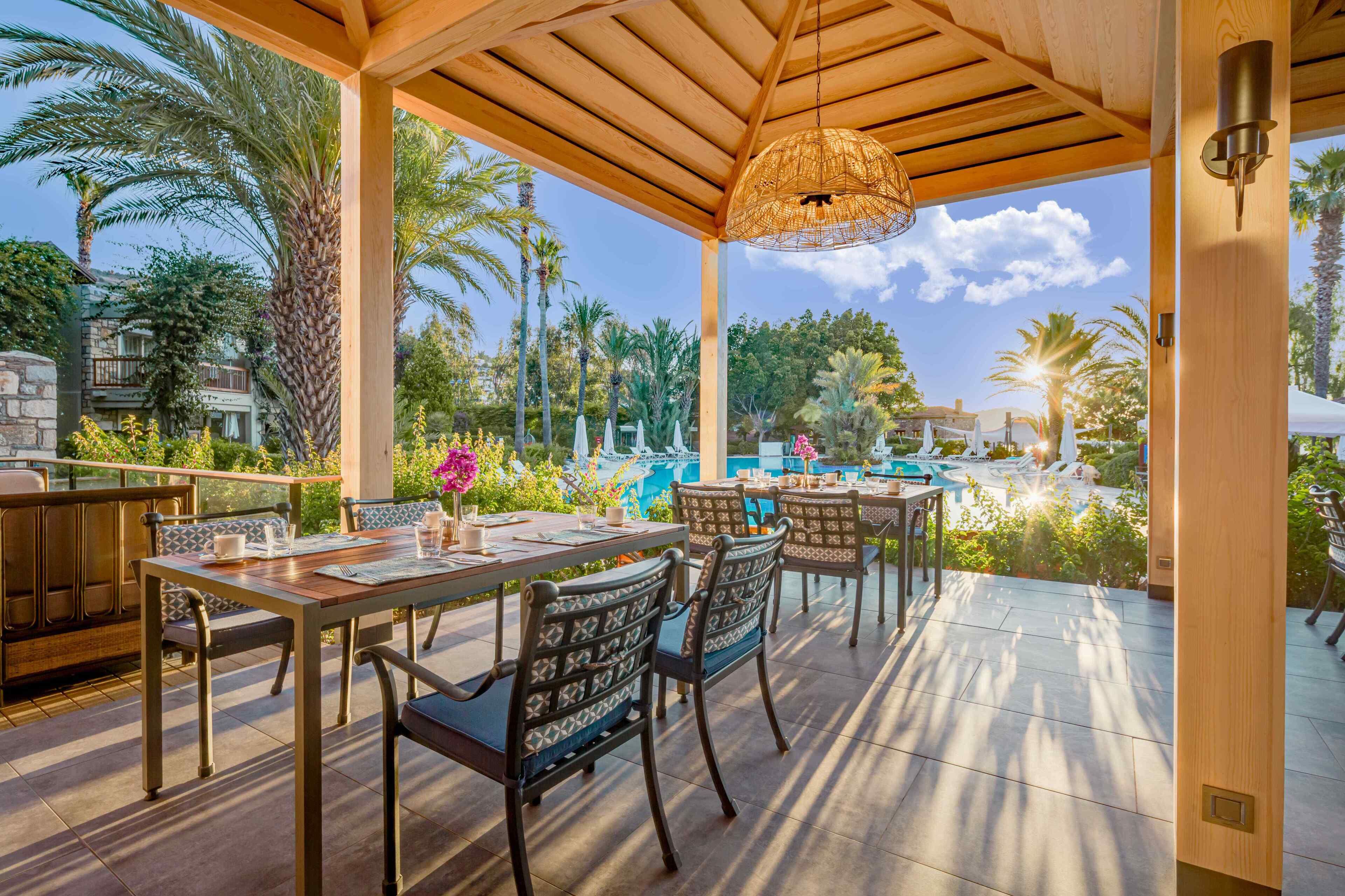 An outdoor patio with wooden tables and chairs, a hanging light fixture, and a view of a pool and palm trees.