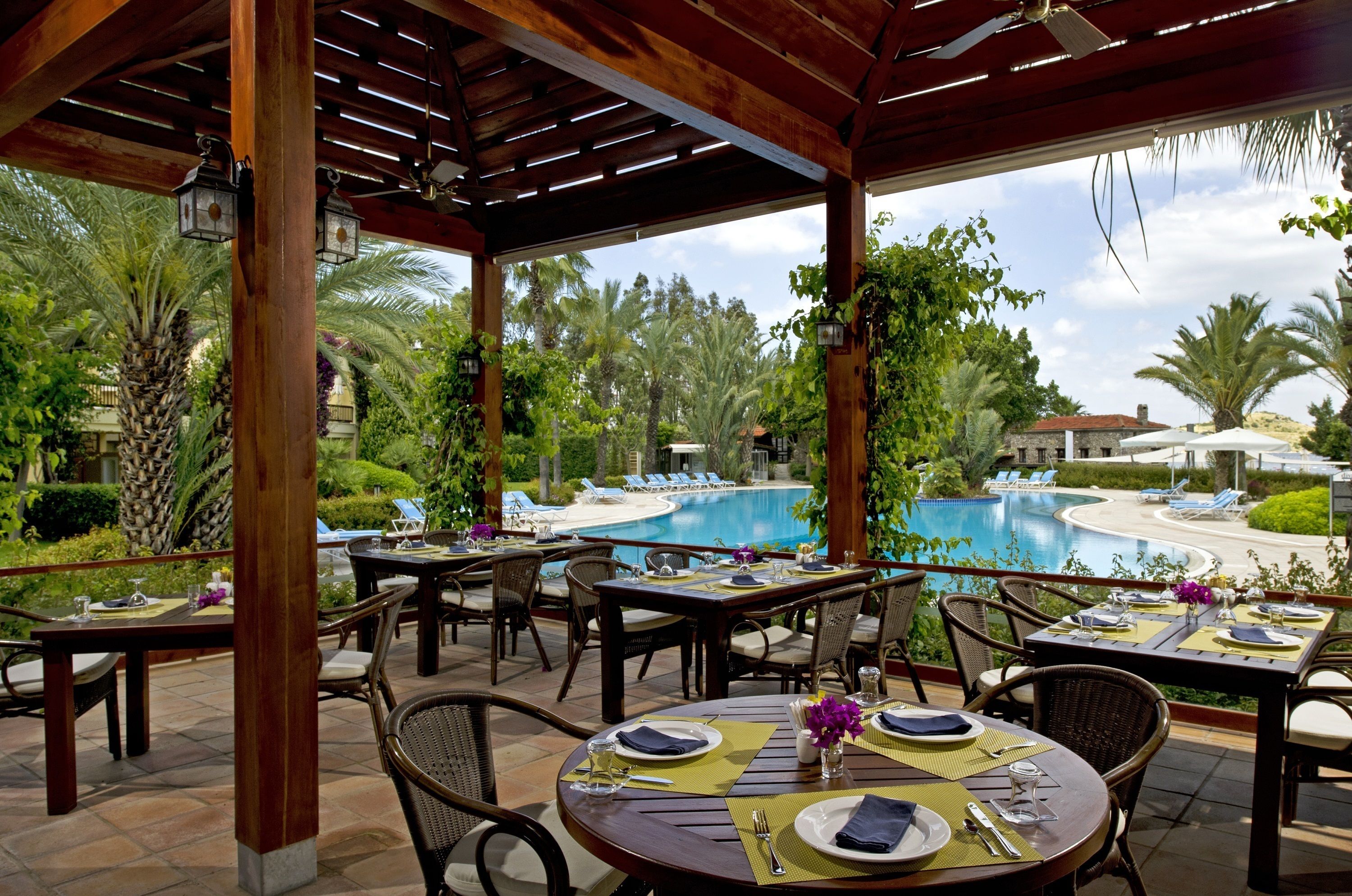 An outdoor dining area with tables set for two, overlooking a pool and lush greenery.
