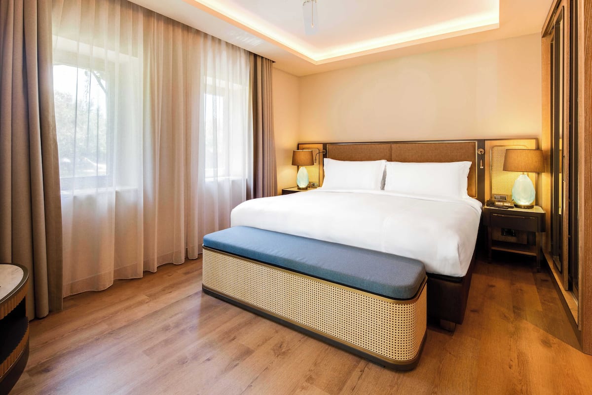 Habitación de hotel con una cama grande, dos lámparas de noche, un banco y vistas al exterior a través de cortinas translúcidas.