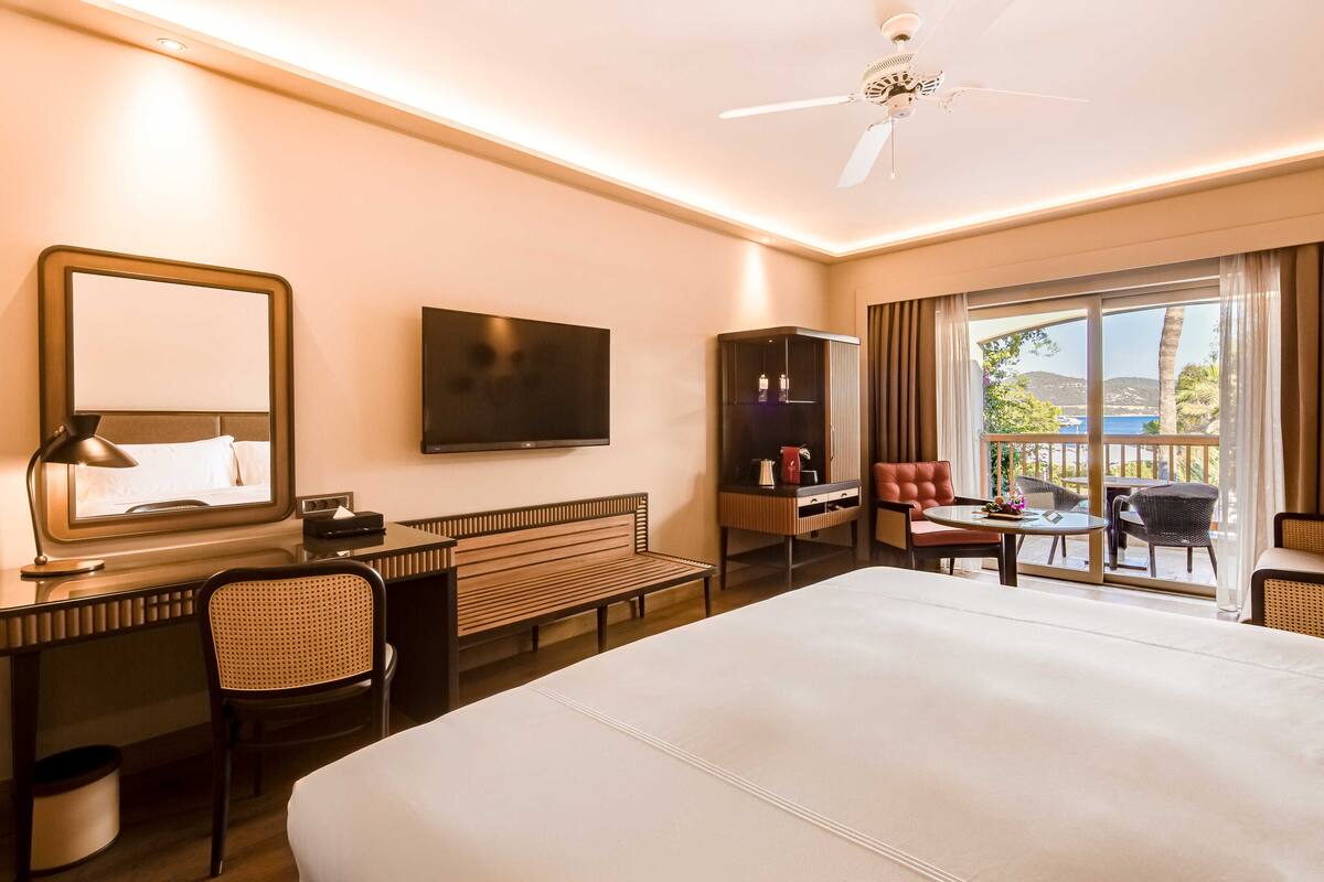Habitación de hotel con una cama grande, un escritorio con silla, televisión, armario y balcón con mobiliario.