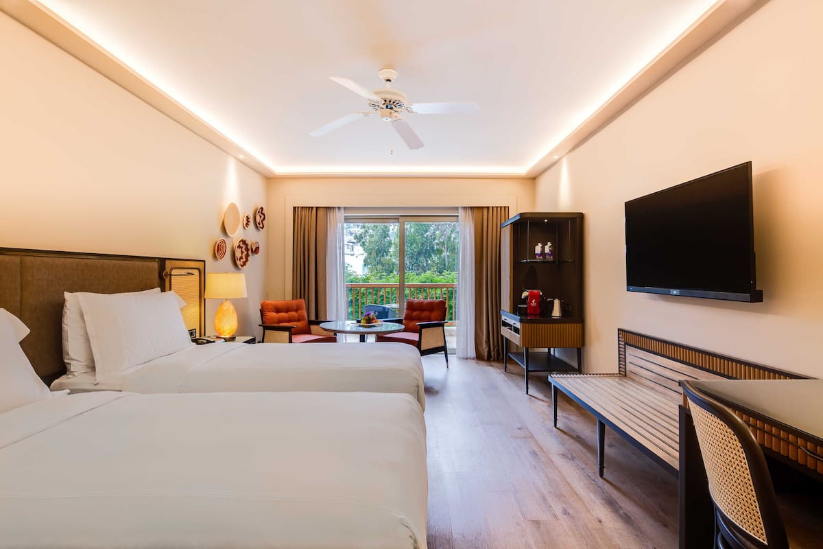 Habitación de hotel con dos camas, televisión, ventilador de techo, balcón con vistas y escritorio con silla.