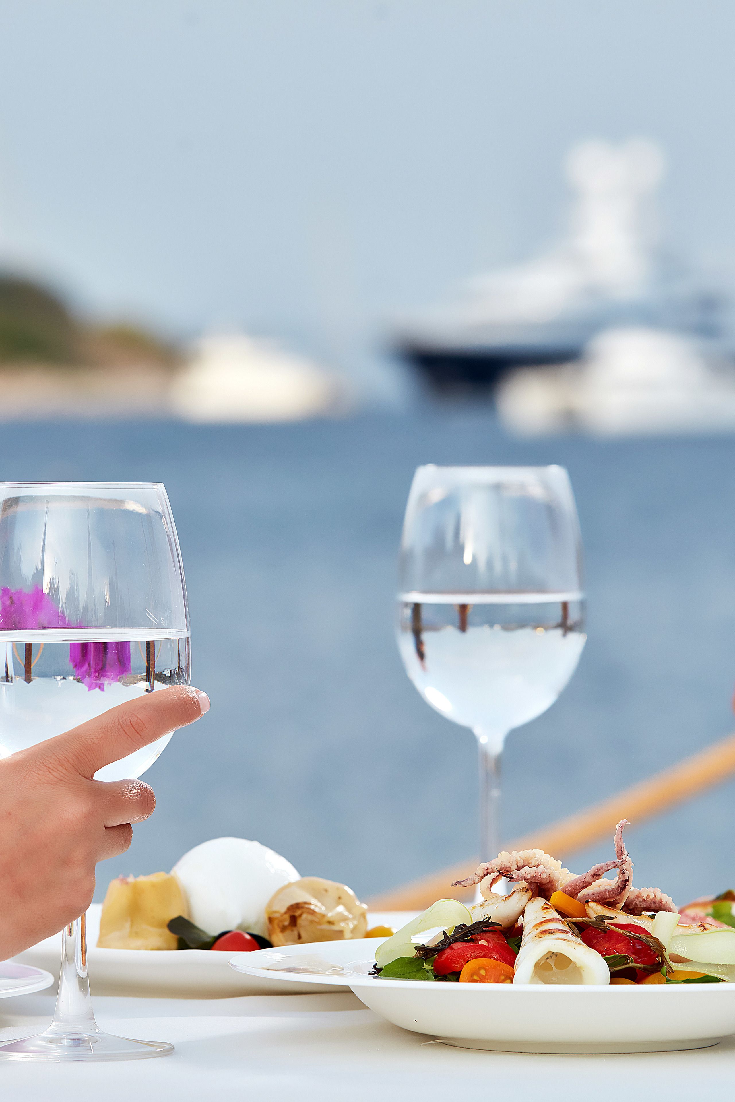 A dining table with a seafood dish and two wine glasses, set against a backdrop of a body of water and a boat.