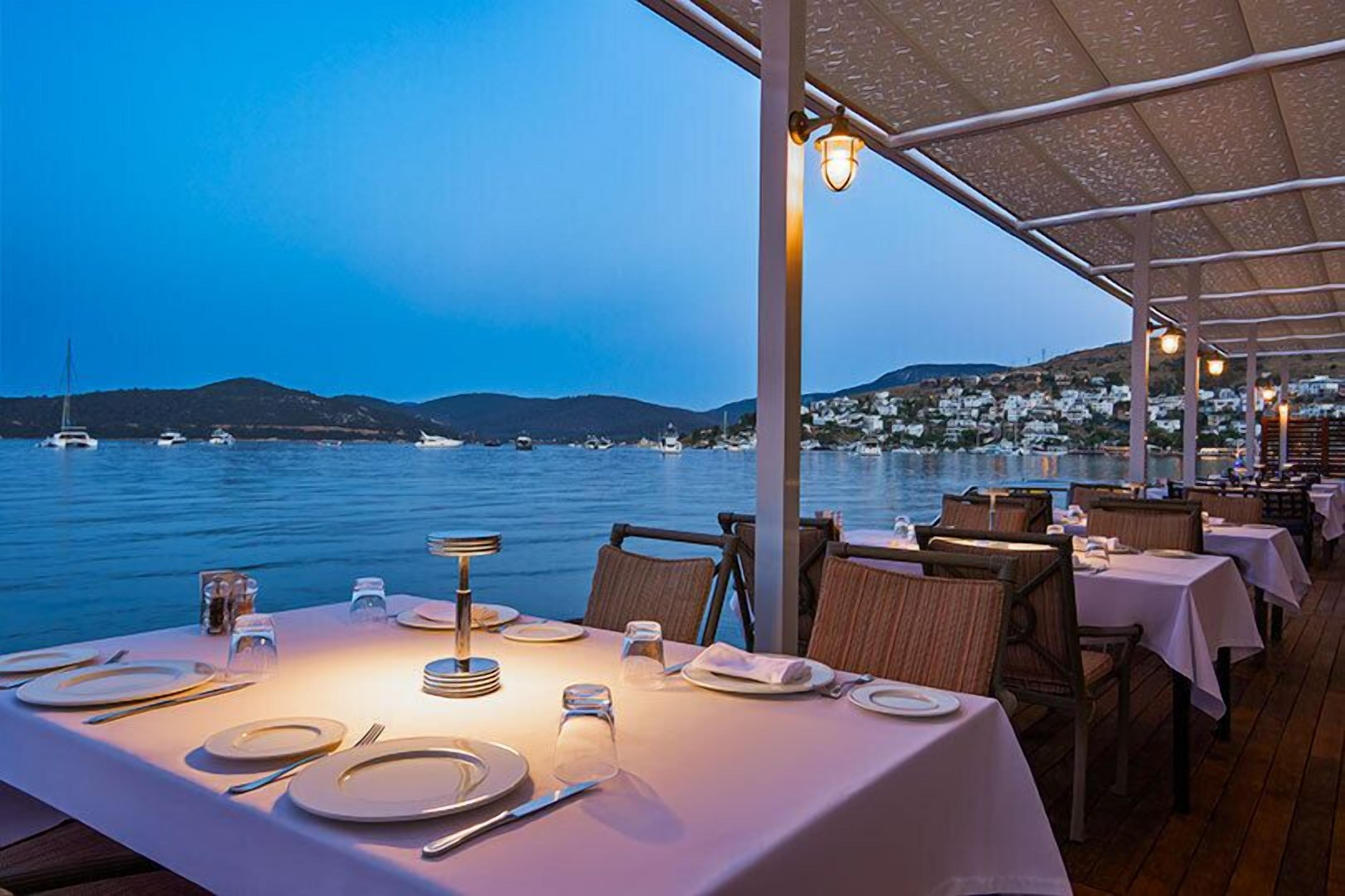 A dining area with tables set for a meal, overlooking a body of water with boats and a hilly landscape in the background.