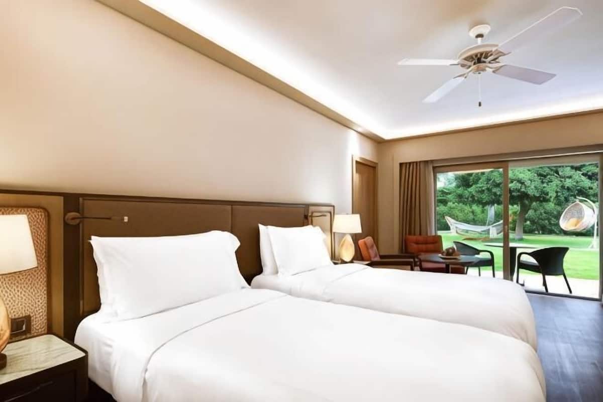 Habitación de hotel con dos camas, un ventilador de techo, una mesita de noche con lámpara, un escritorio con silla y vistas al exterior.