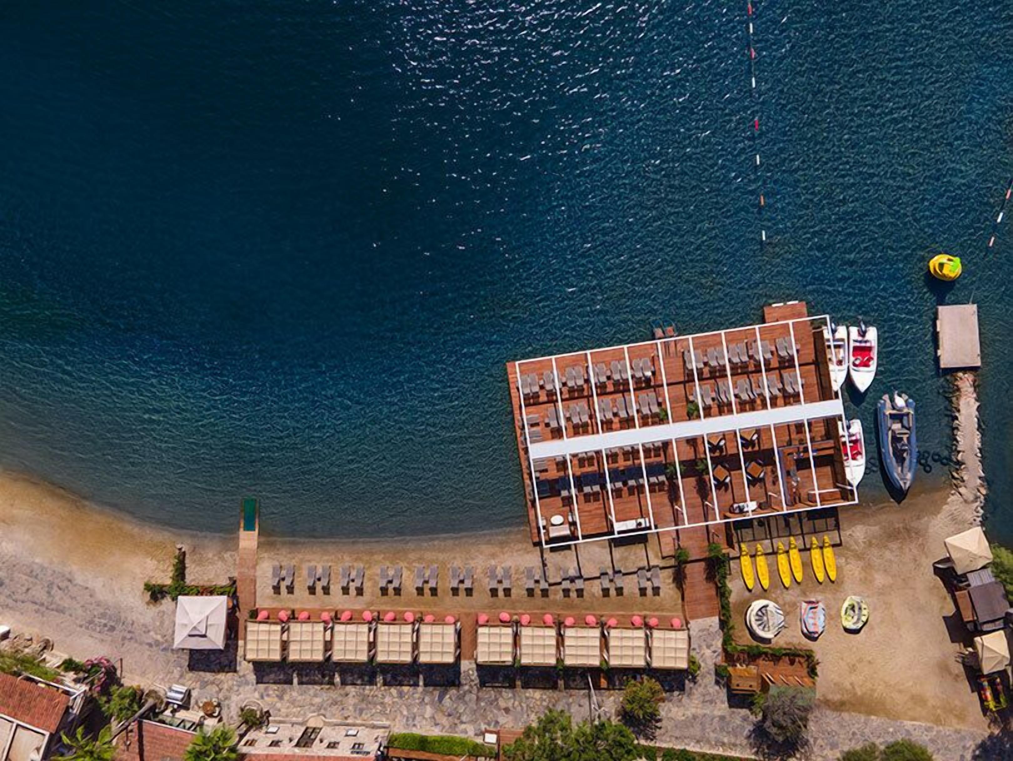 An aerial view of a coastal hotel with a long dining terrace, a pool area, and several boats.