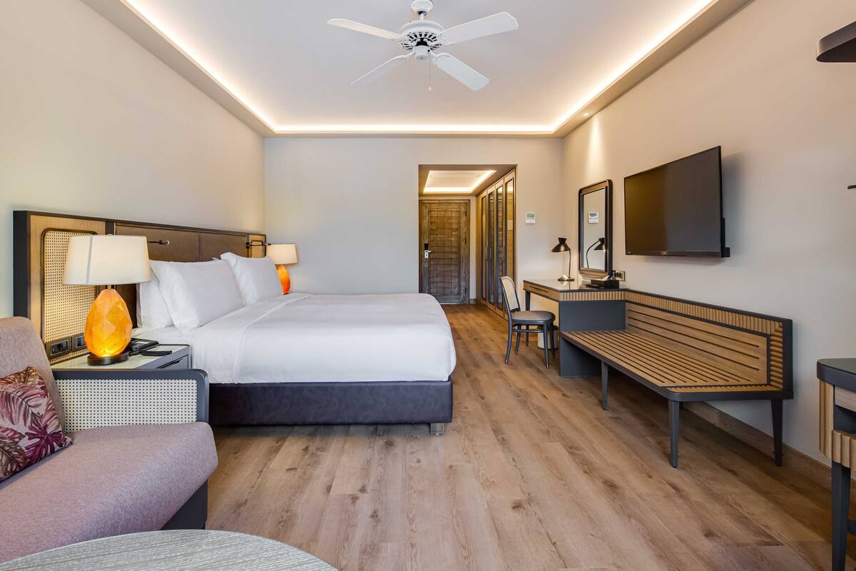 Una habitación de hotel moderna con cama, mesitas de noche, escritorio, silla, banco, ventilador de techo y televisor montado en la pared.