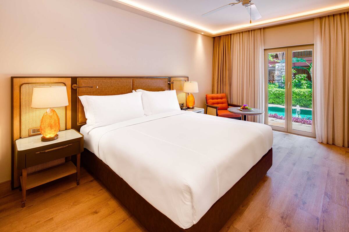 Habitación de hotel con una cama grande, una mesita de noche con lámpara, una silla y vista a un jardín por una puerta corrediza.