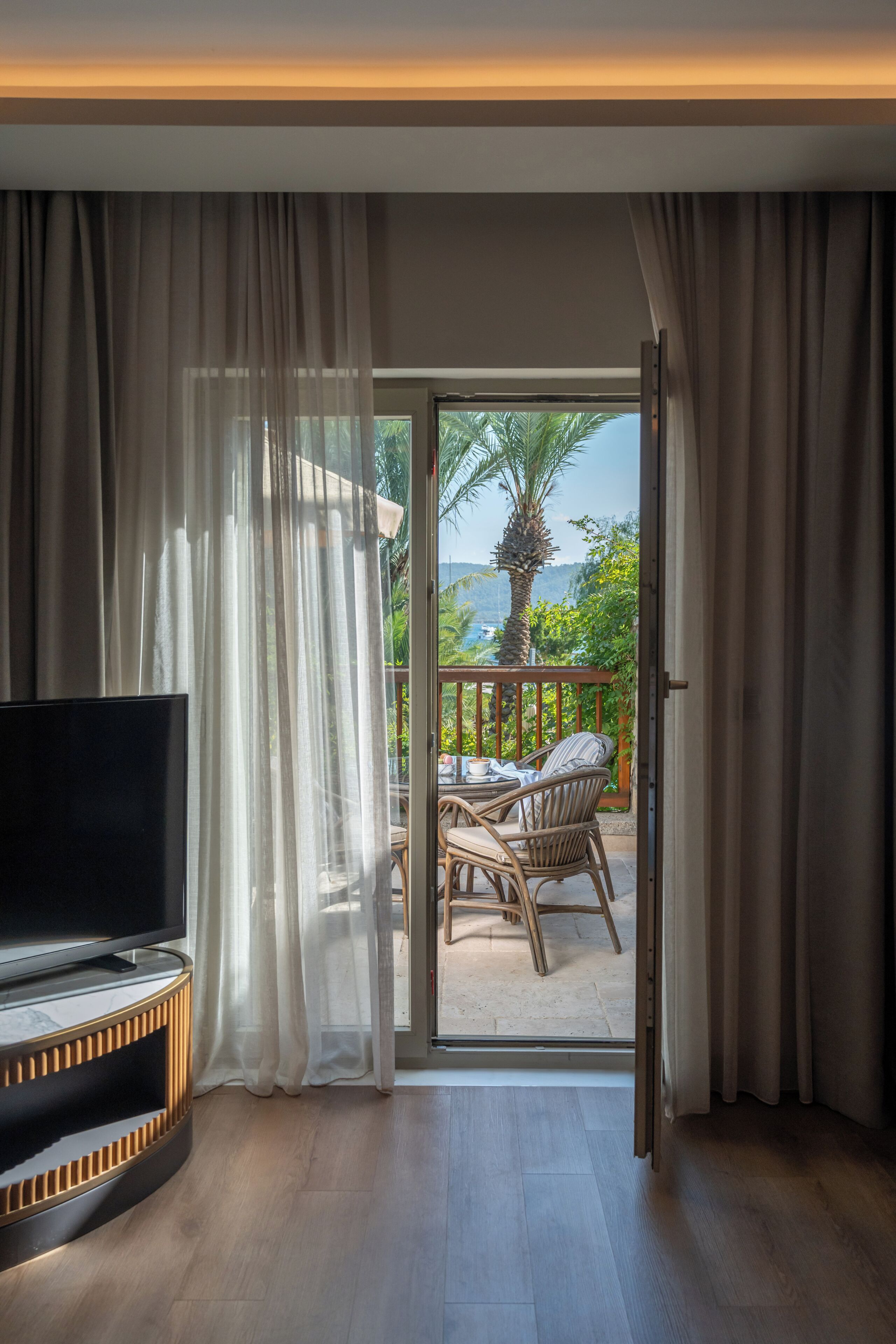 Habitación con televisión, piso de madera, balcón con mesa y sillas, y vistas al exterior.