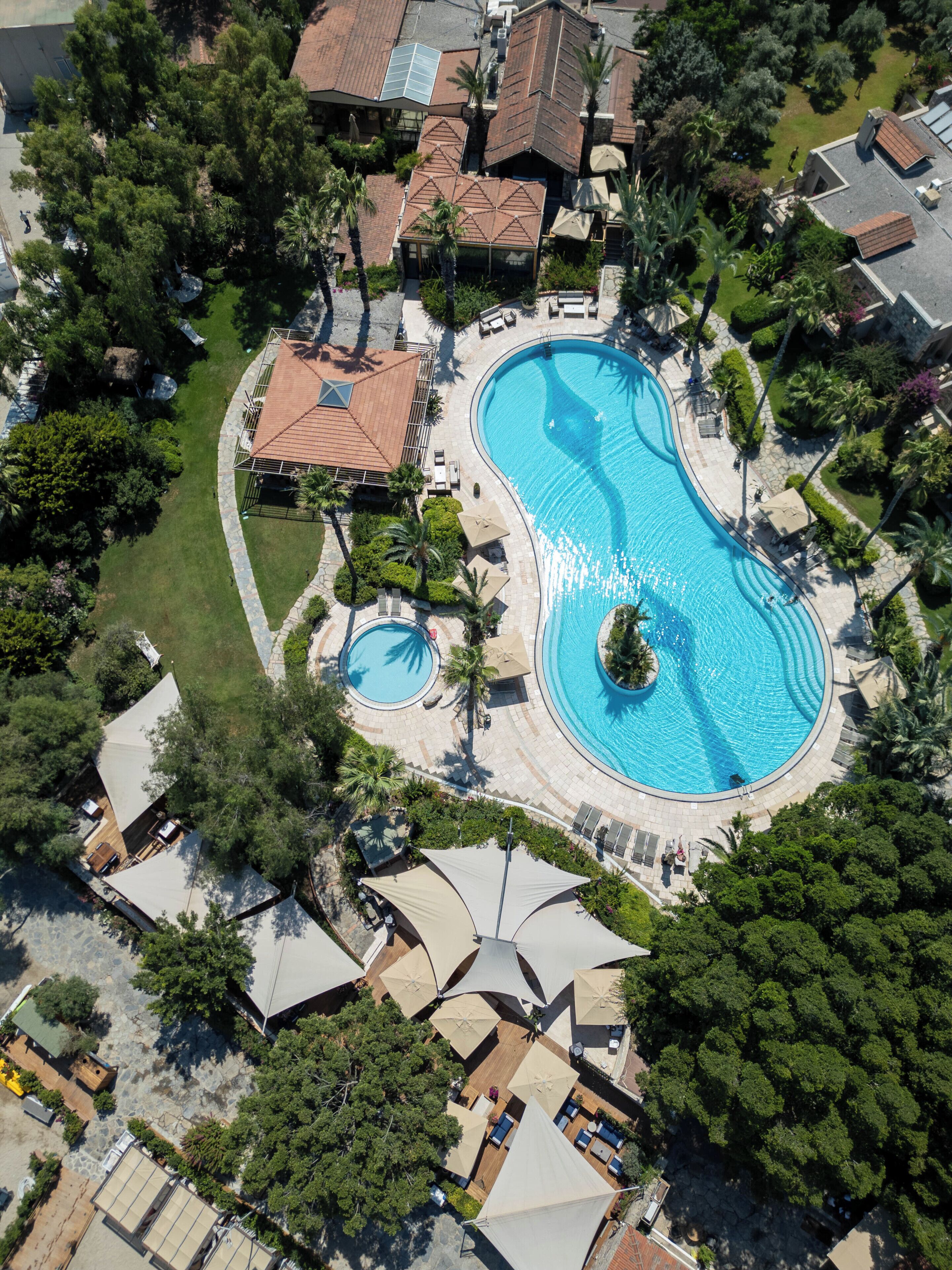 Vue aérienne d’un complexe hôtelier doté d’une grande piscine, de plusieurs espaces de détente et d’une végétation luxuriante.