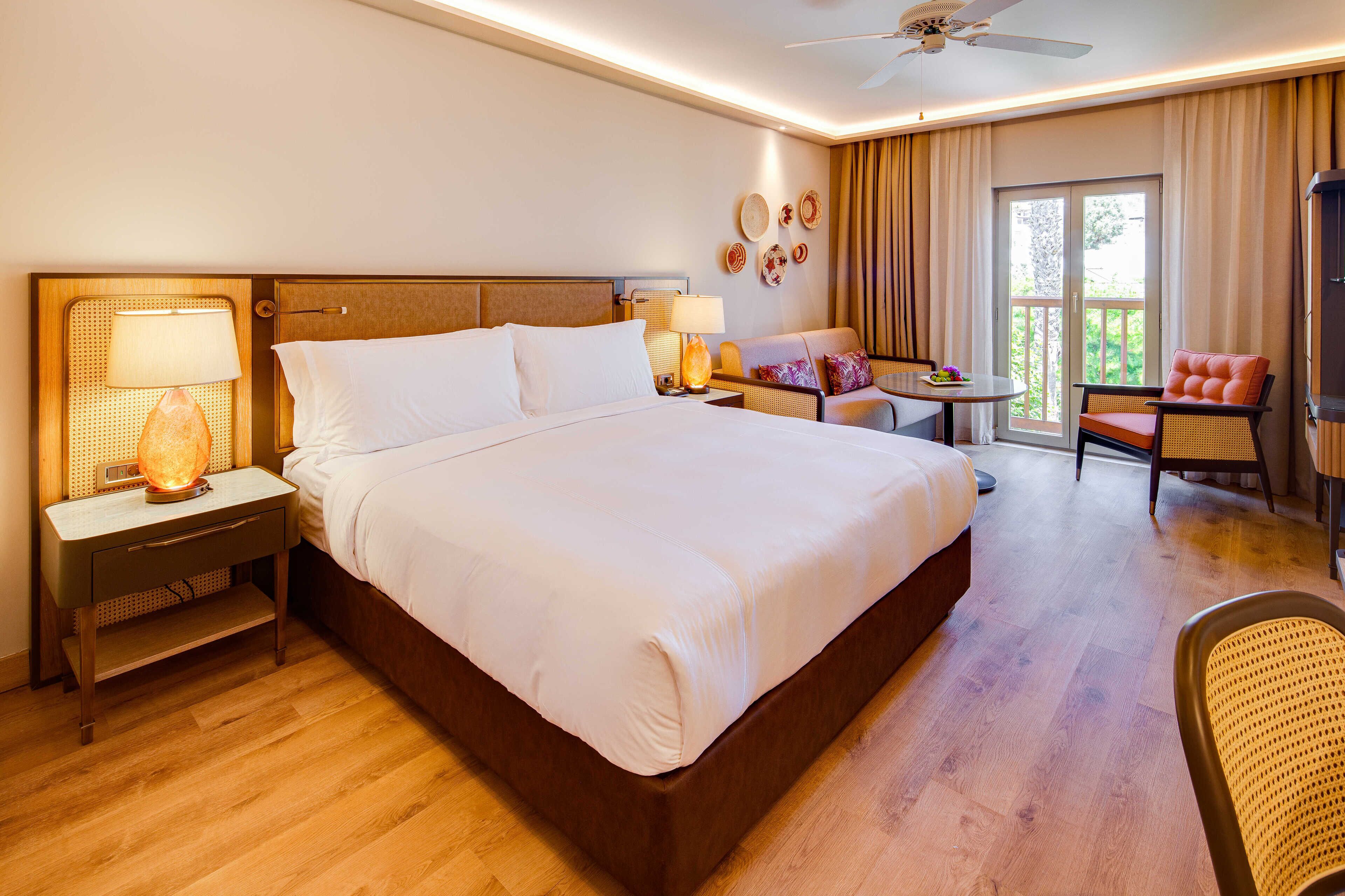 A hotel room with a large bed, a desk with a lamp, a chair, and a balcony with curtains.