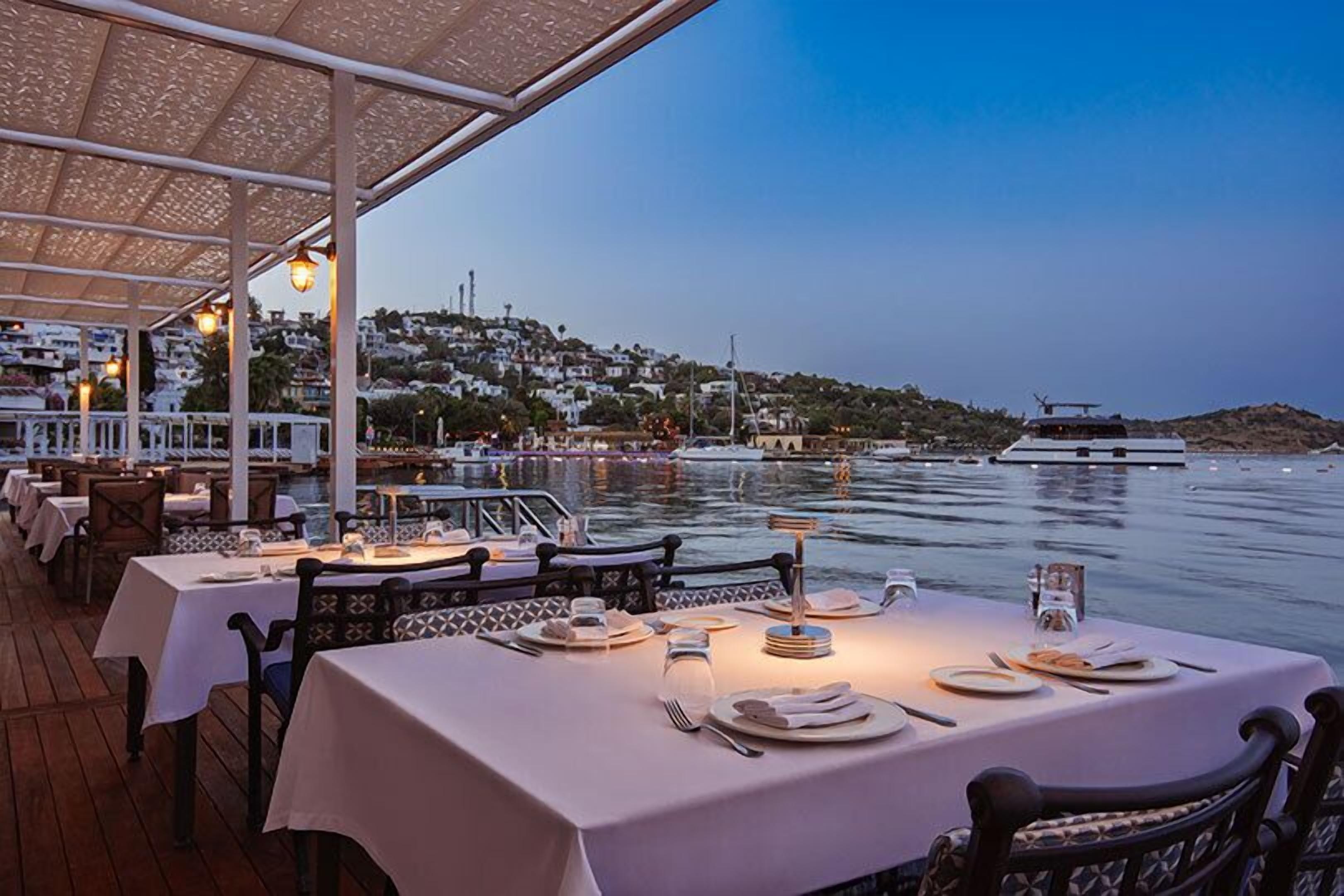 Un restaurant situé au bord de l’eau, avec des tables dressées pour le dîner, offrant une vue sur une étendue d’eau où naviguent des bateaux, et sur un paysage vallonné en arrière-plan.