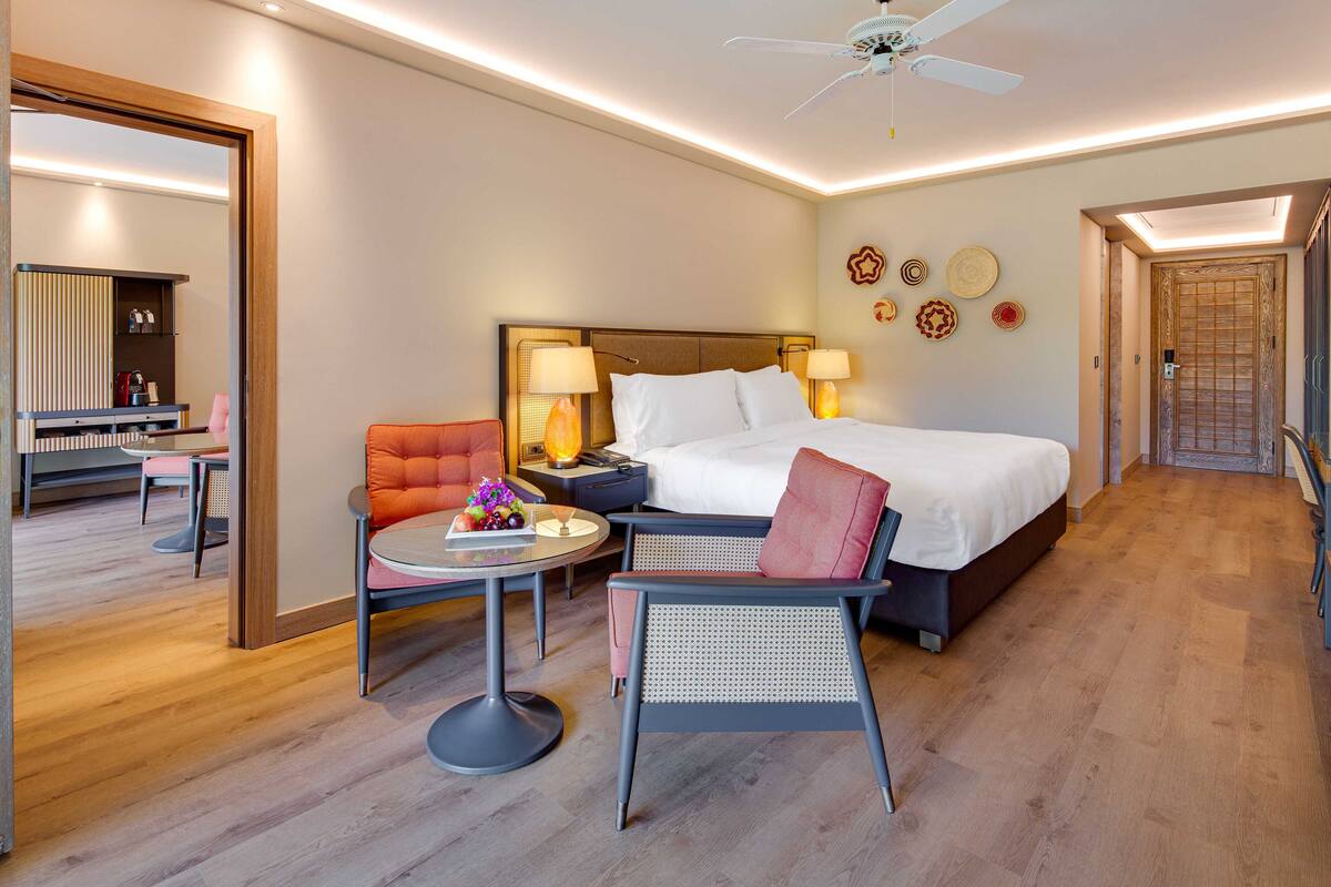 Una habitación de hotel con una cama grande, dos sillas, una mesita y un ventilador de techo.