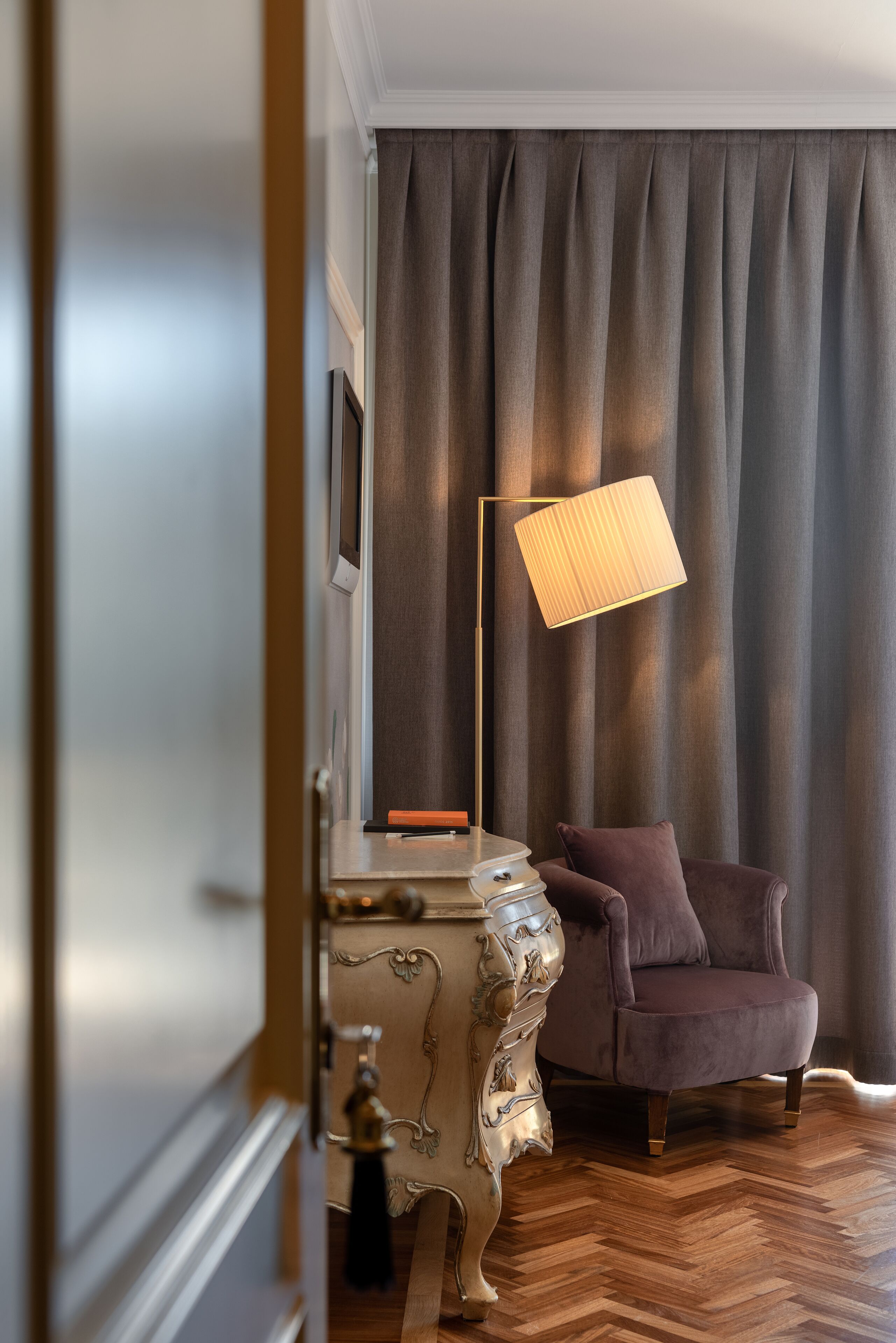 Una stanza con pavimento in legno a spina di pesce, tenda scura, lampada da terra moderna, poltrona imbottita e un tavolino decorativo.